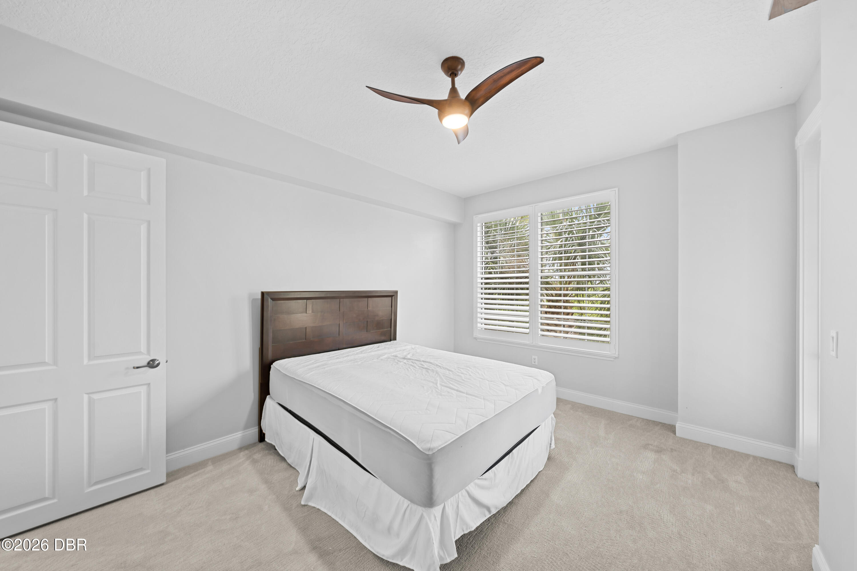 Bedroom with a white bed, dark wood headboard, shuttered window, and sculptural ceiling fan.