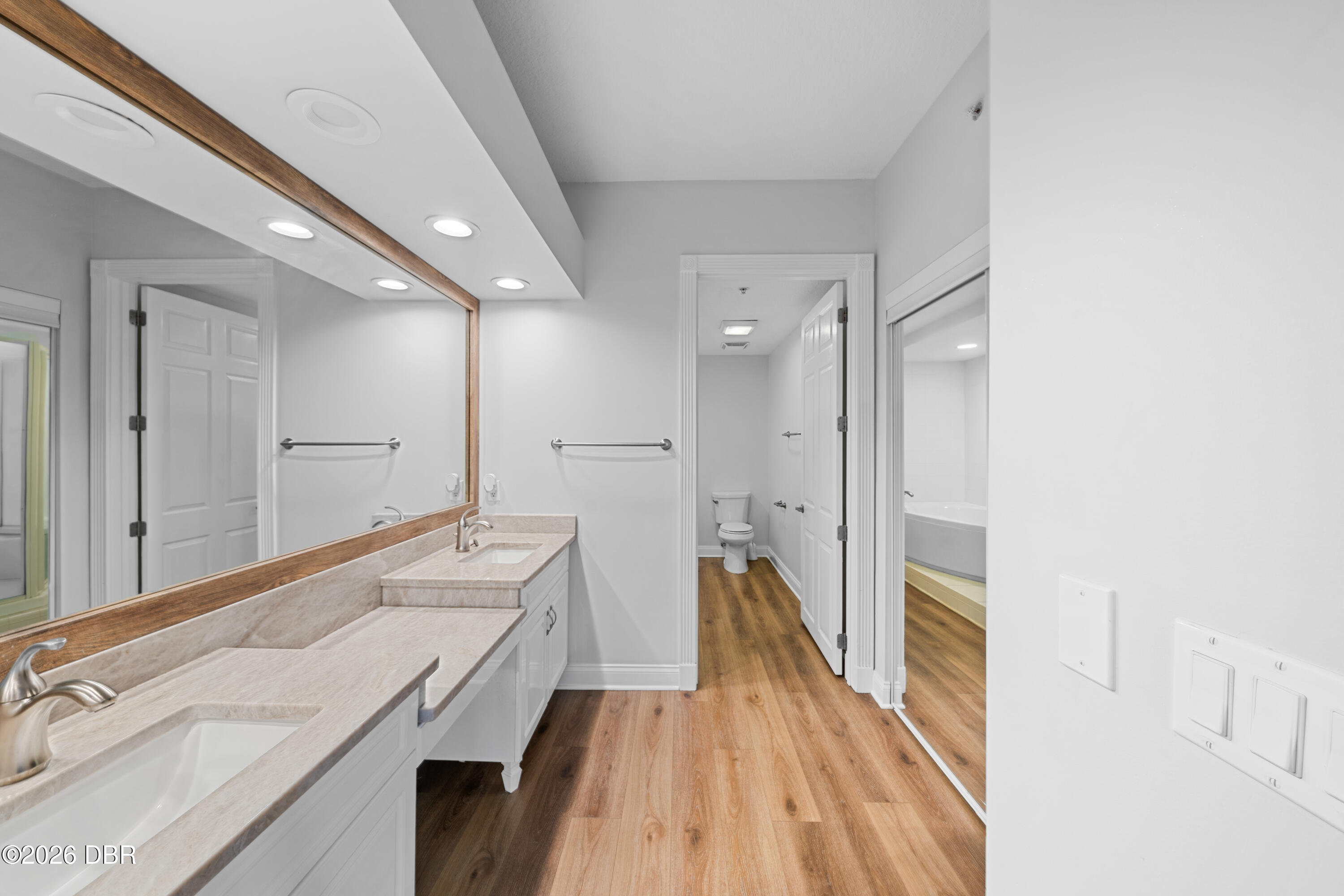 Bathroom with a long stone vanity, wood-framed mirror, and wood-look flooring leading to a separate toilet area.
