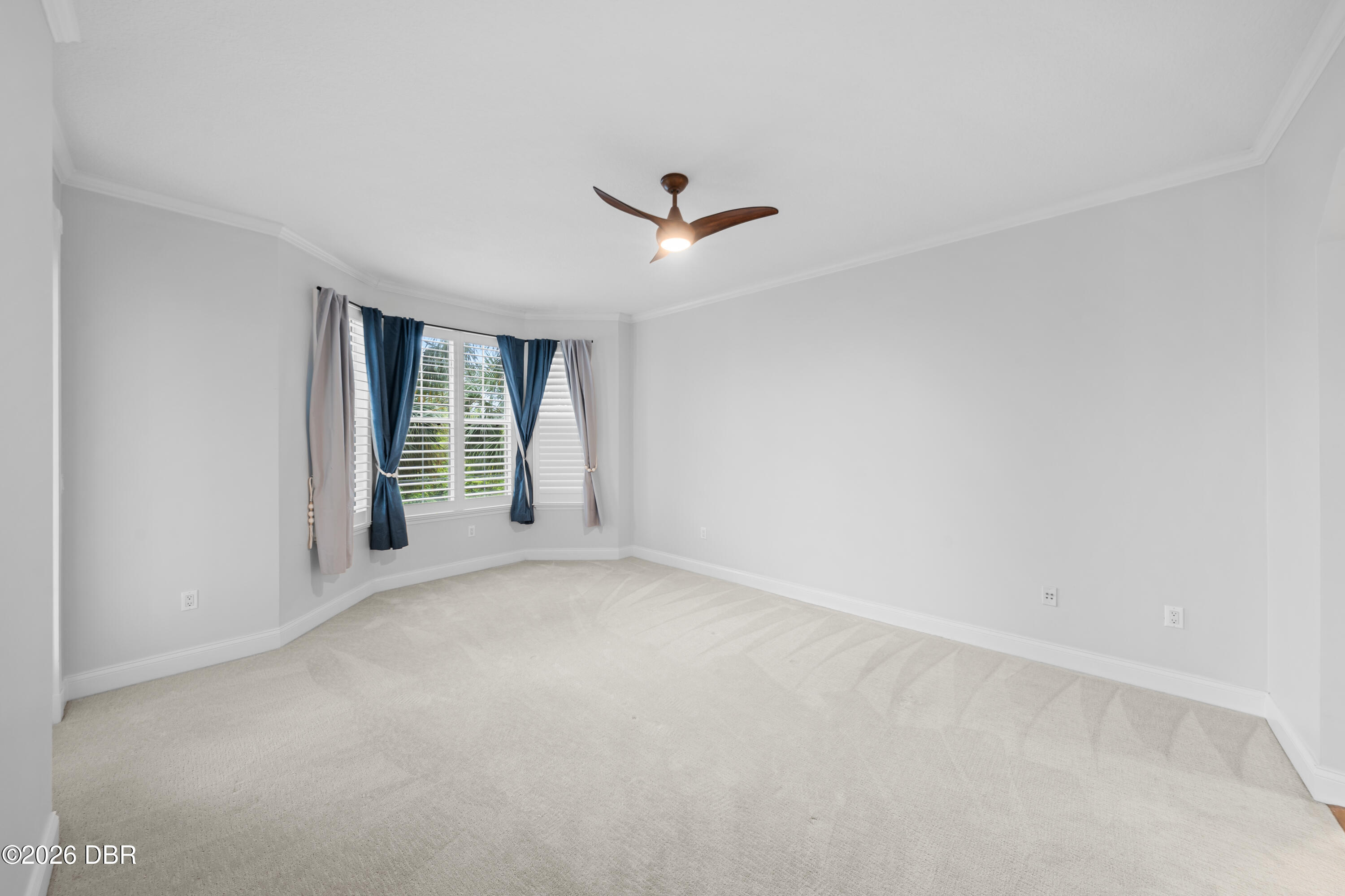 Empty bedroom with a shuttered bay window, blue curtains, carpet, and a wood ceiling fan.