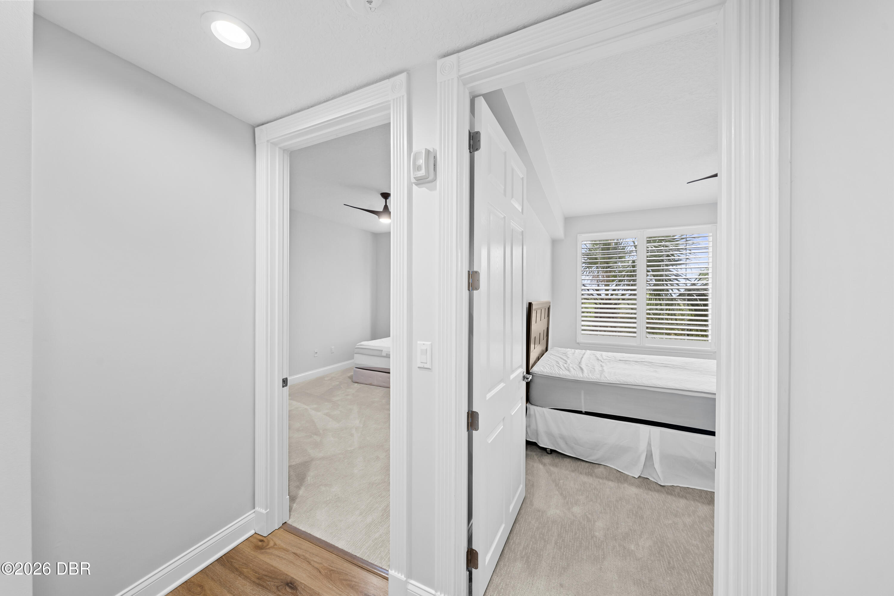 Bedroom with a bed beneath plantation shutters, viewed through an open doorway to a second bedroom.