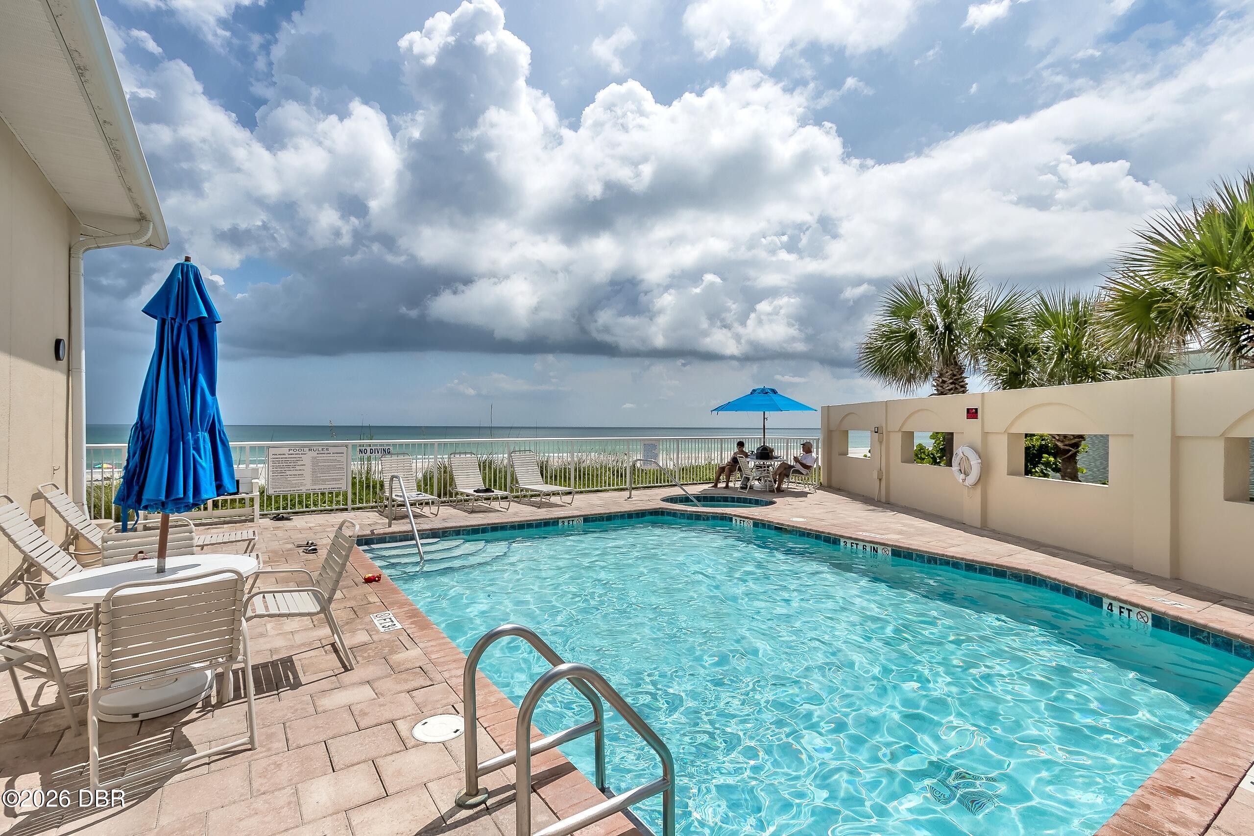 Outdoor pool overlooking the ocean, with blue umbrellas and palm trees.