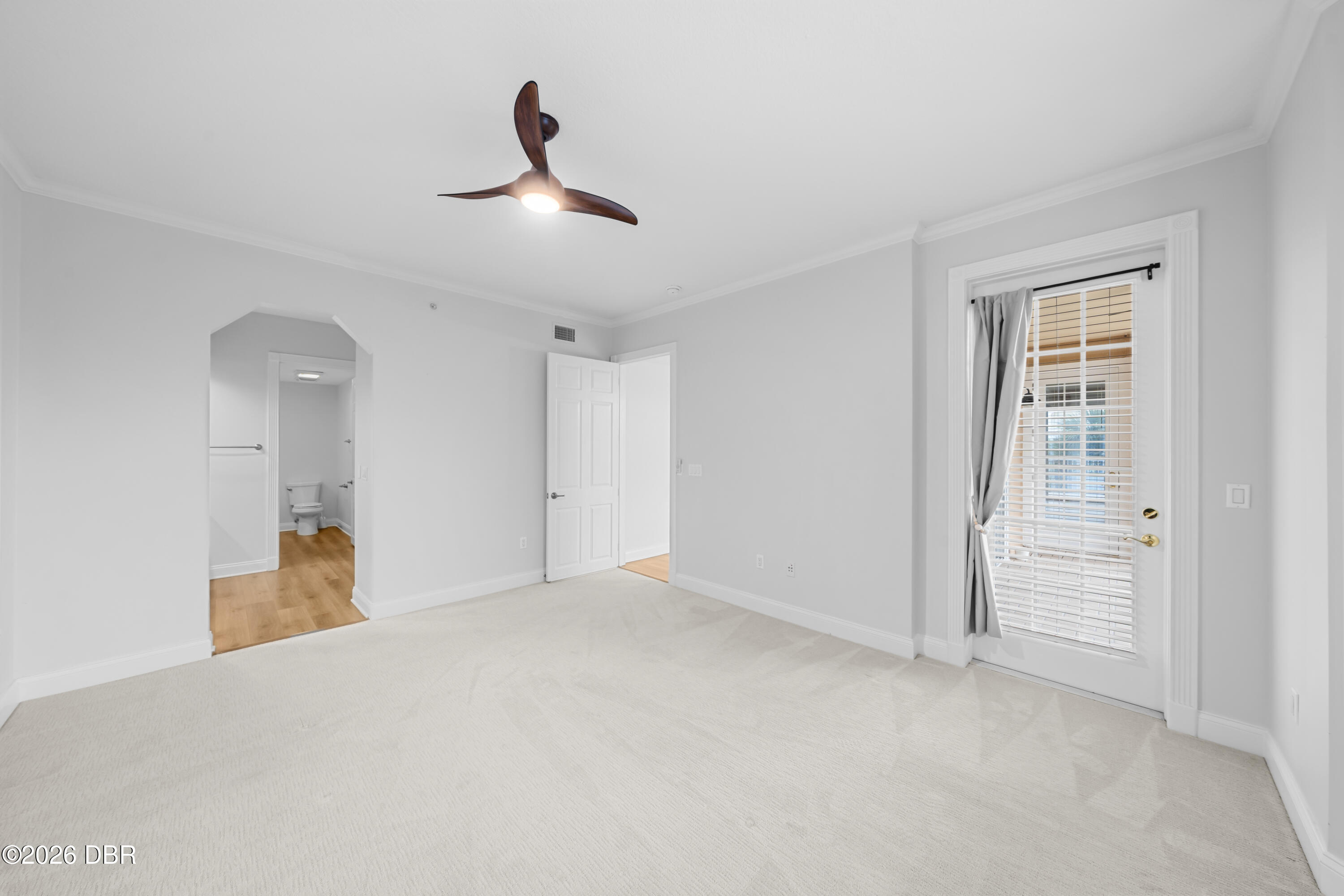 Empty carpeted room with a ceiling fan, curtained glass door, and open doorway to a bathroom.