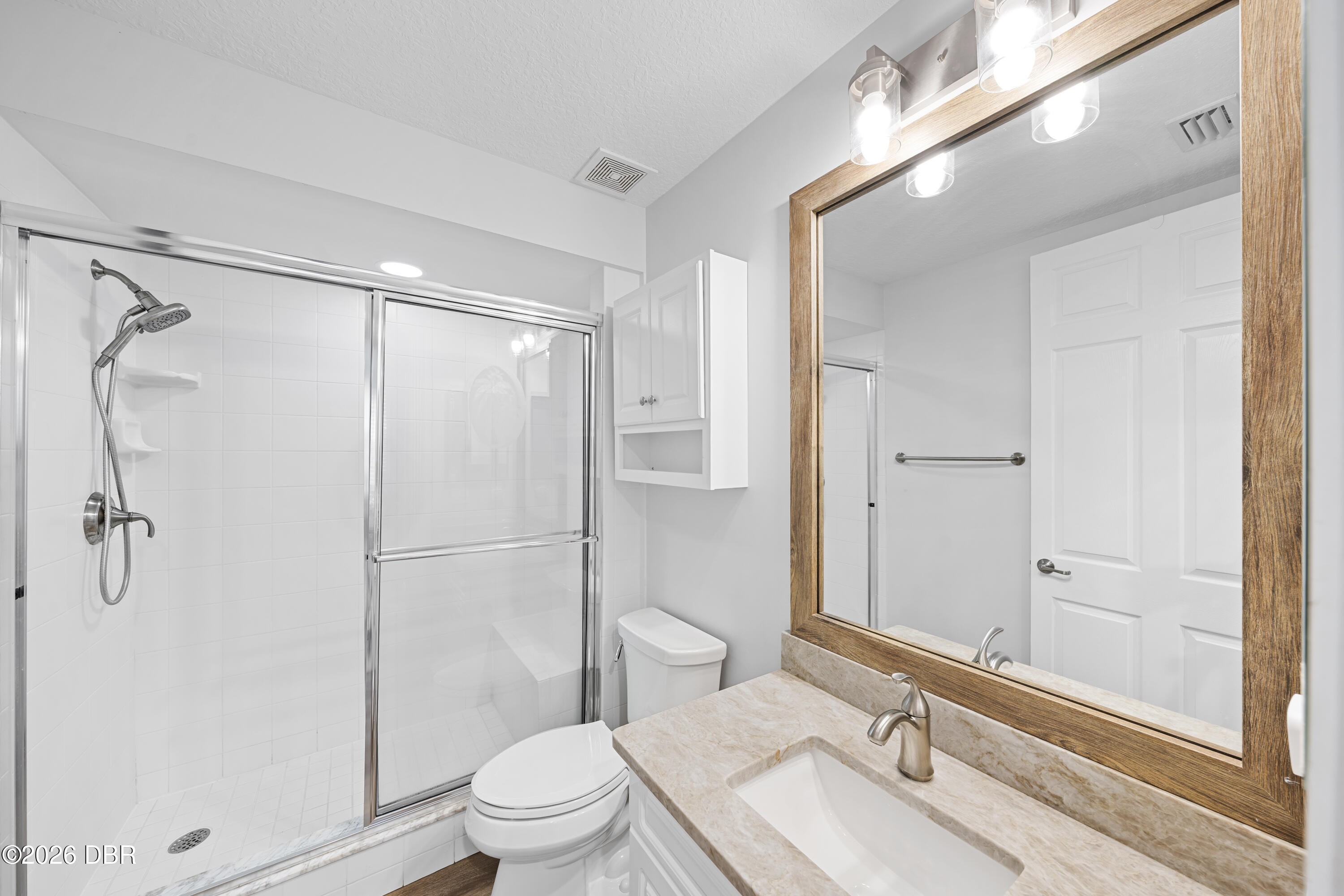 Bathroom with a glass-enclosed tiled shower, toilet, and stone-look vanity beneath a wood-framed mirror.