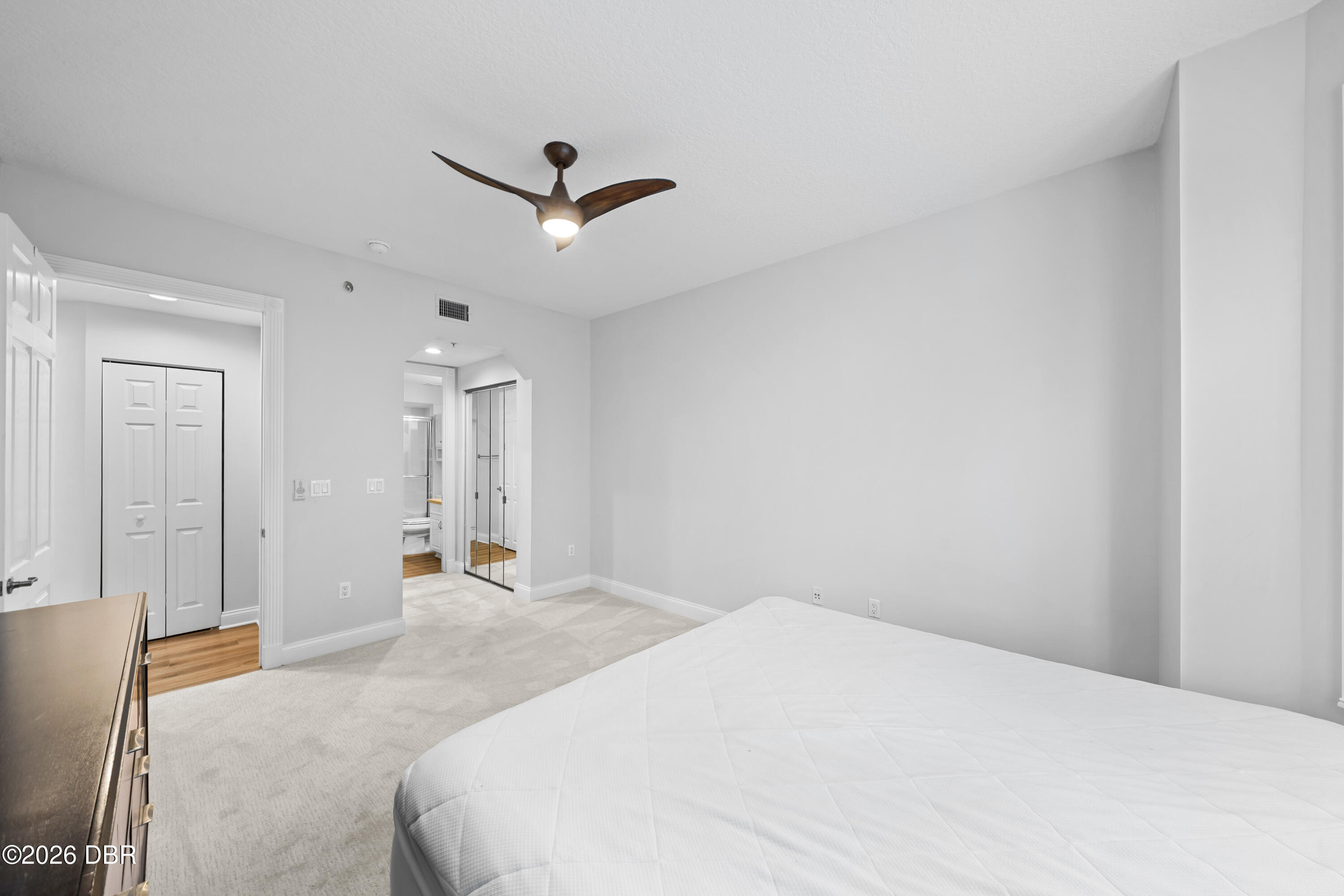 Bedroom with a white bed, dark wood dresser, and ceiling fan beside open closet and bathroom doorways.