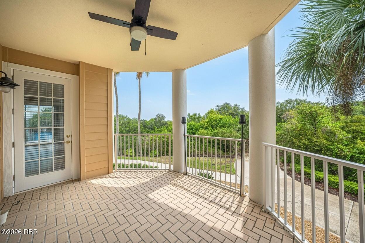 Covered tiled balcony with a ceiling fan and white railing overlooking green trees.