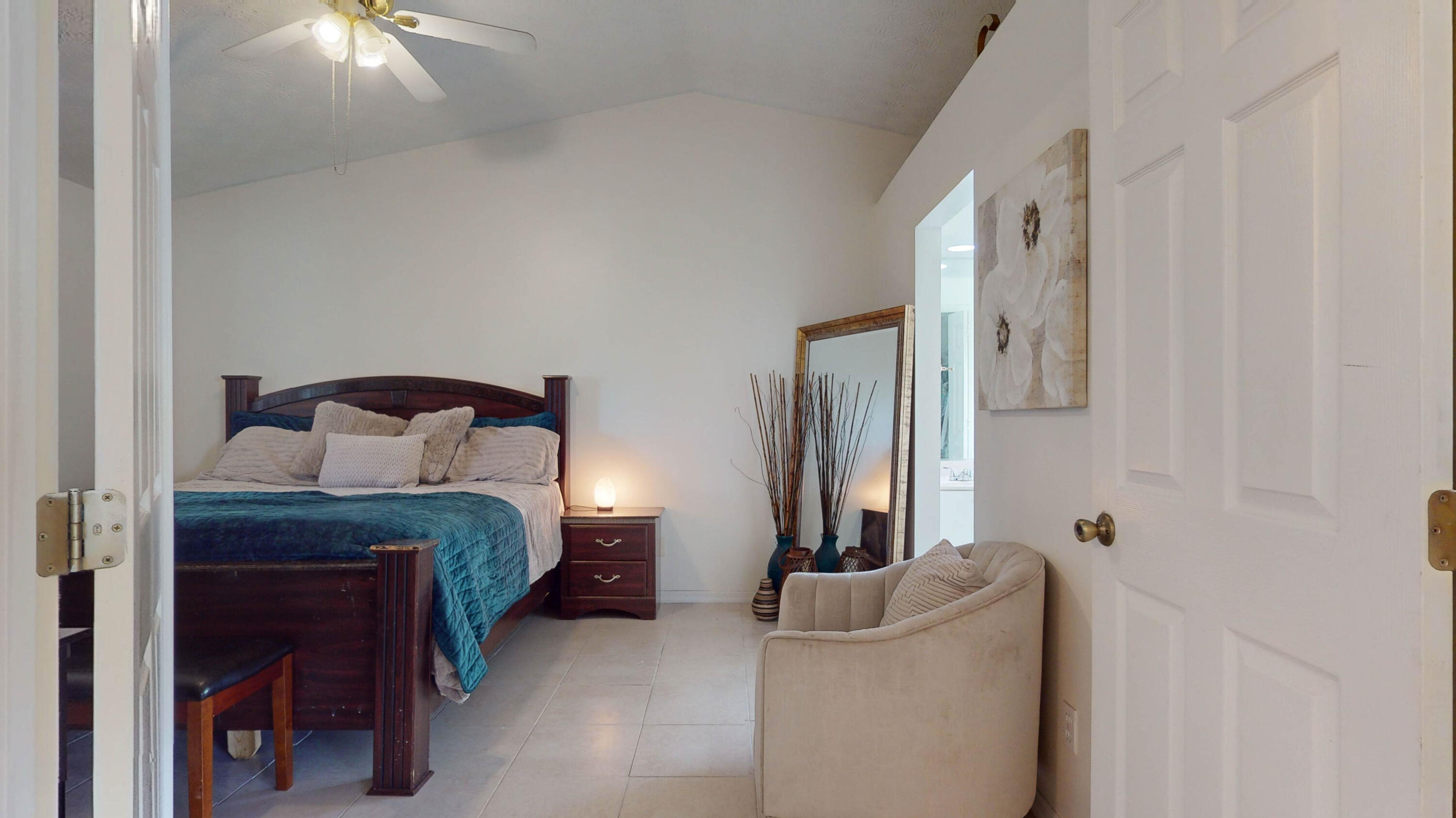 Furnished bedroom with a dark wood bed, teal bedding, beige chair, and tiled floor.
