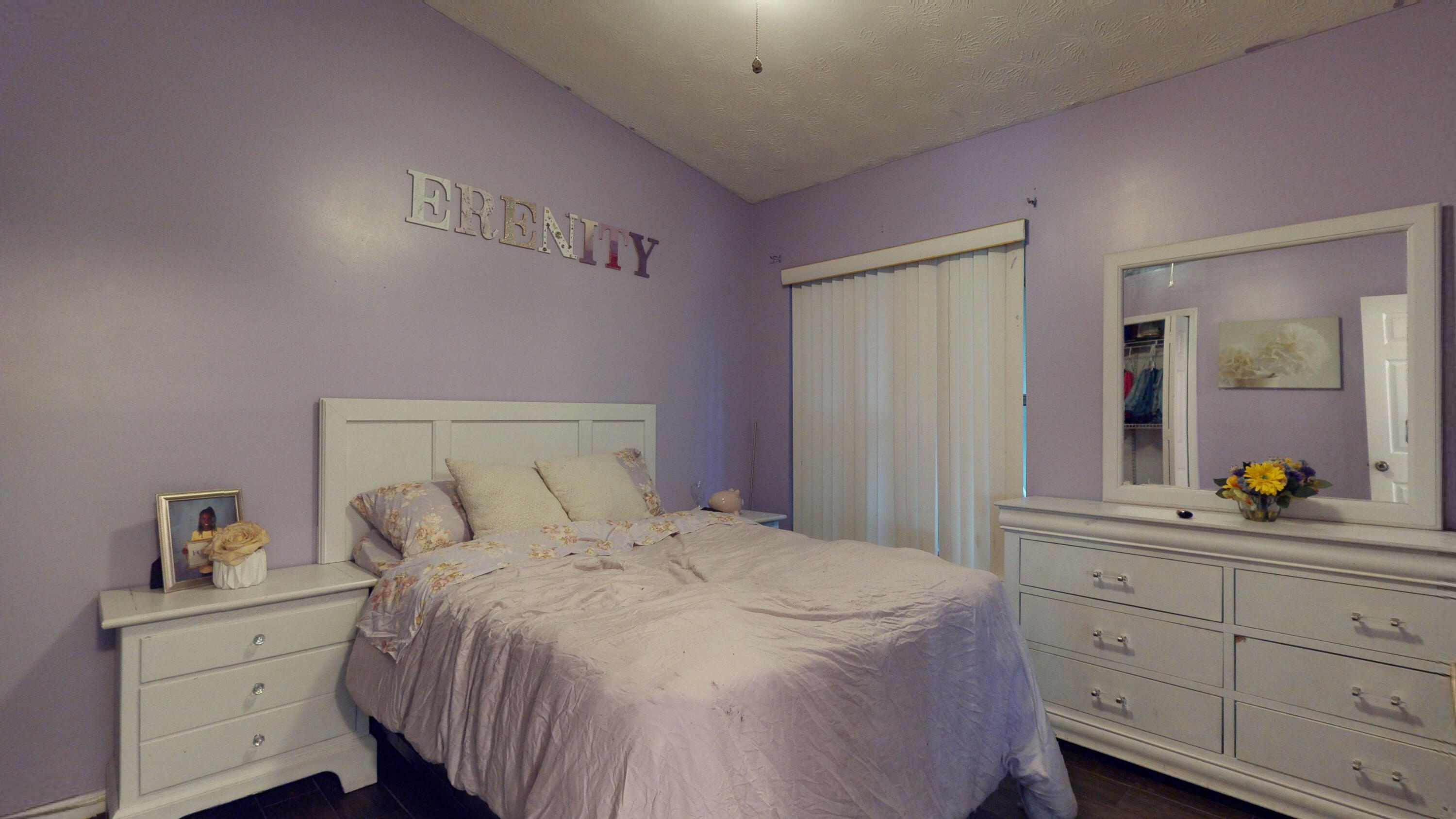 Bedroom with lavender walls, floral bedding, white furniture, and vertical blinds.