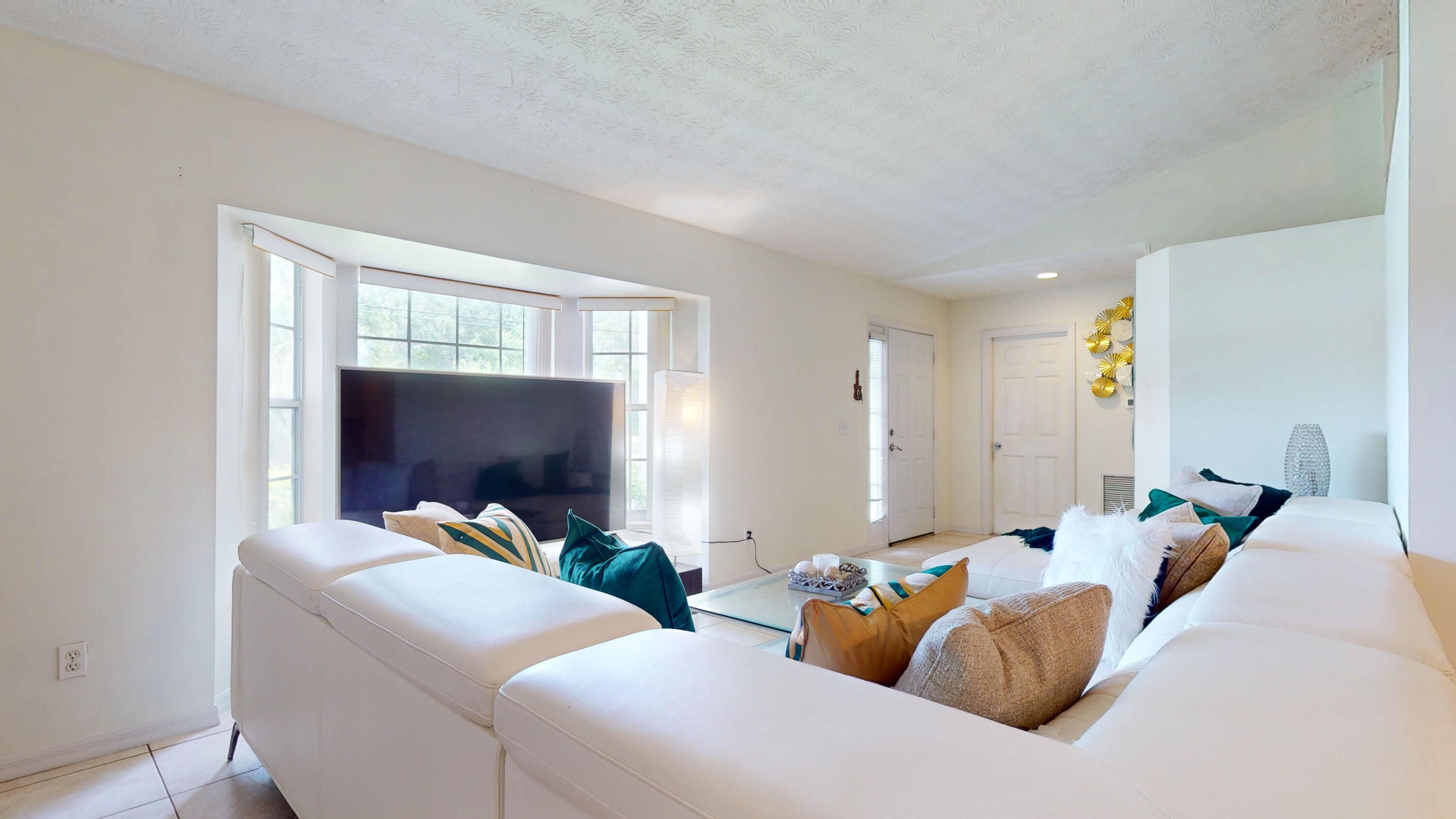 Furnished living room with a white sectional, teal and gold pillows, and bay windows behind a large television.