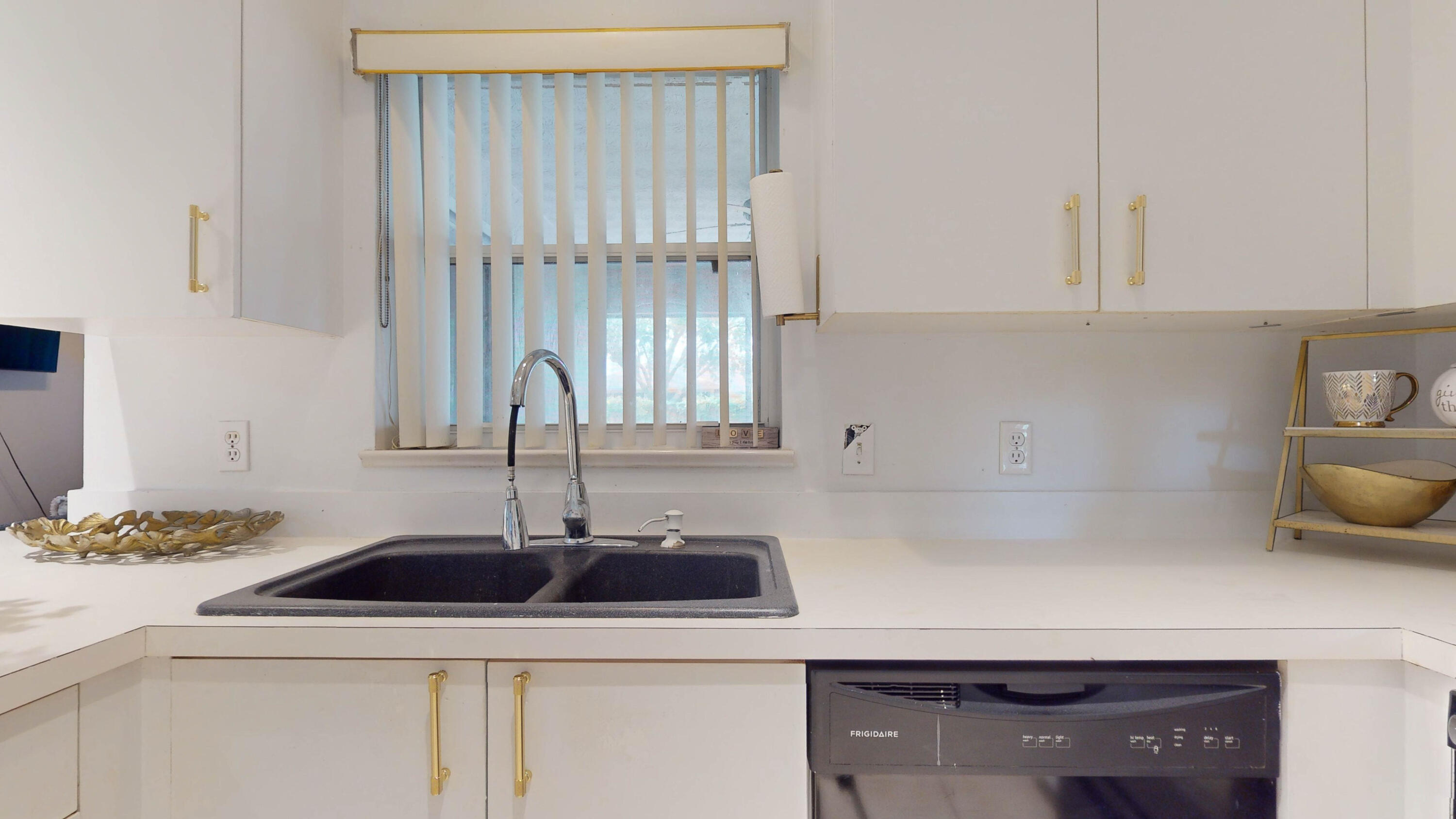 Kitchen sink beneath a window, with white cabinets, gold-tone hardware, and a dishwasher.