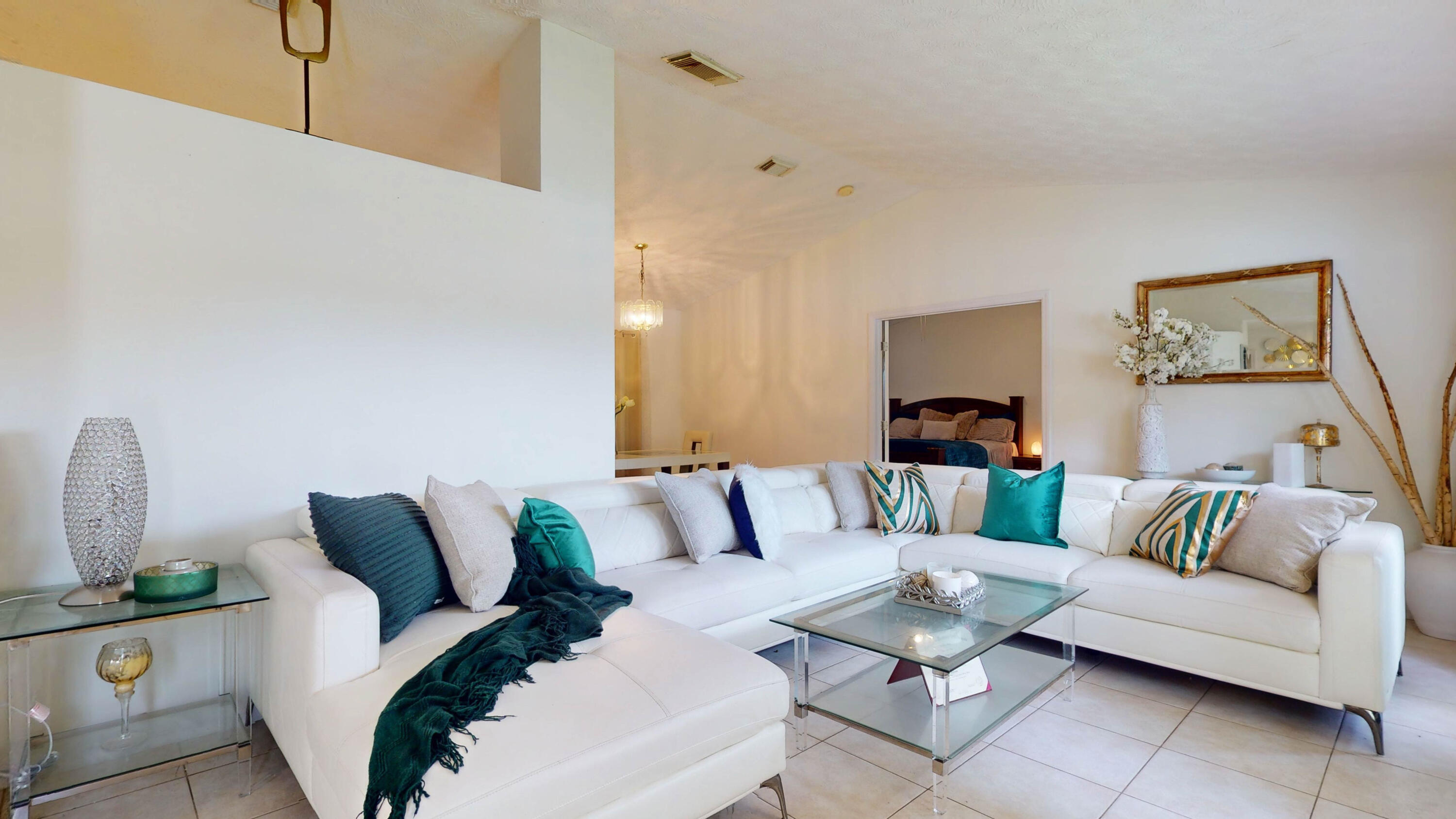 Living room with a white sectional sofa and glass coffee table, with a bedroom visible through the doorway.