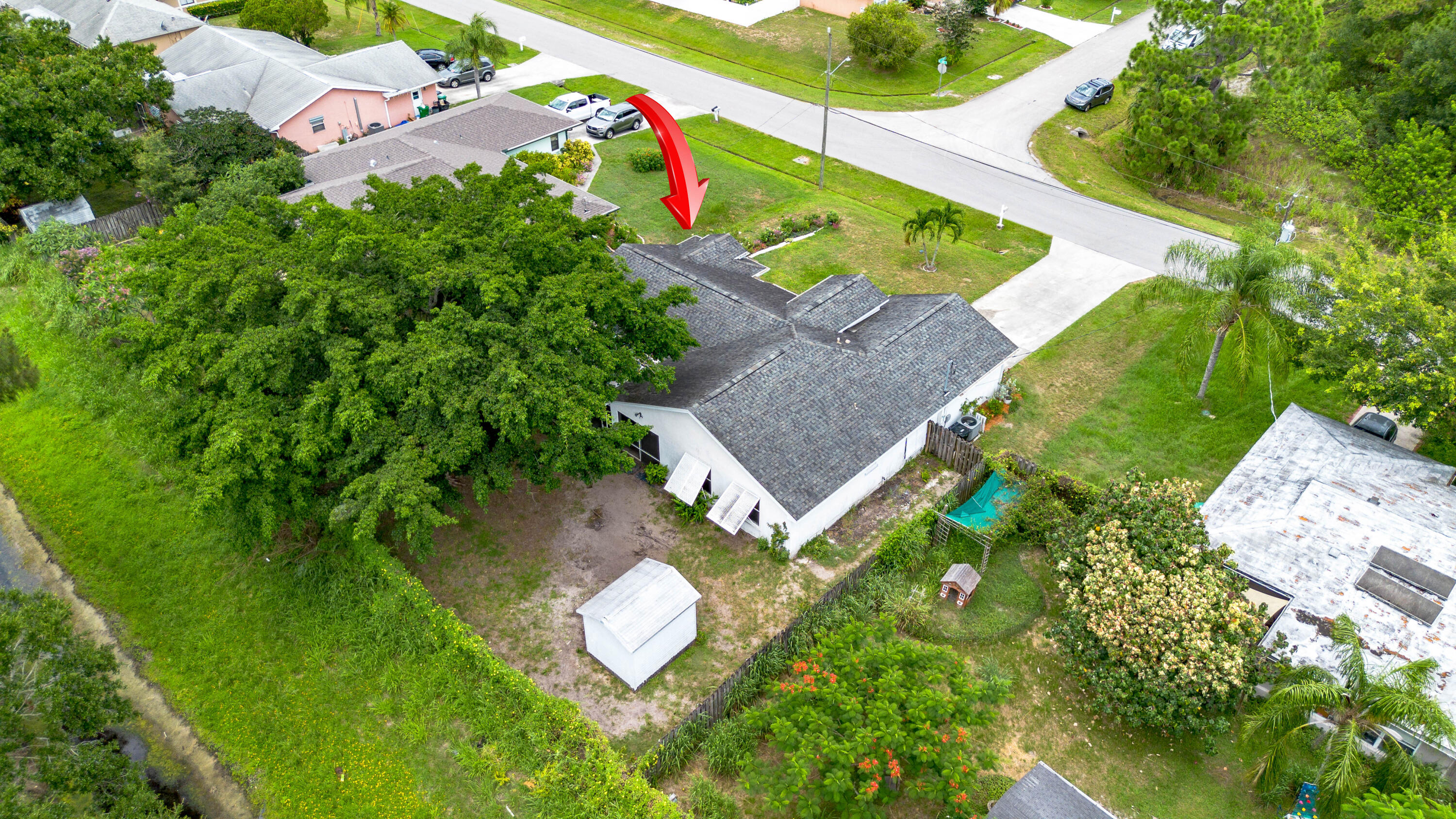 Aerial view of a single-story home, with a red arrow marking the property beside a large tree and white shed.