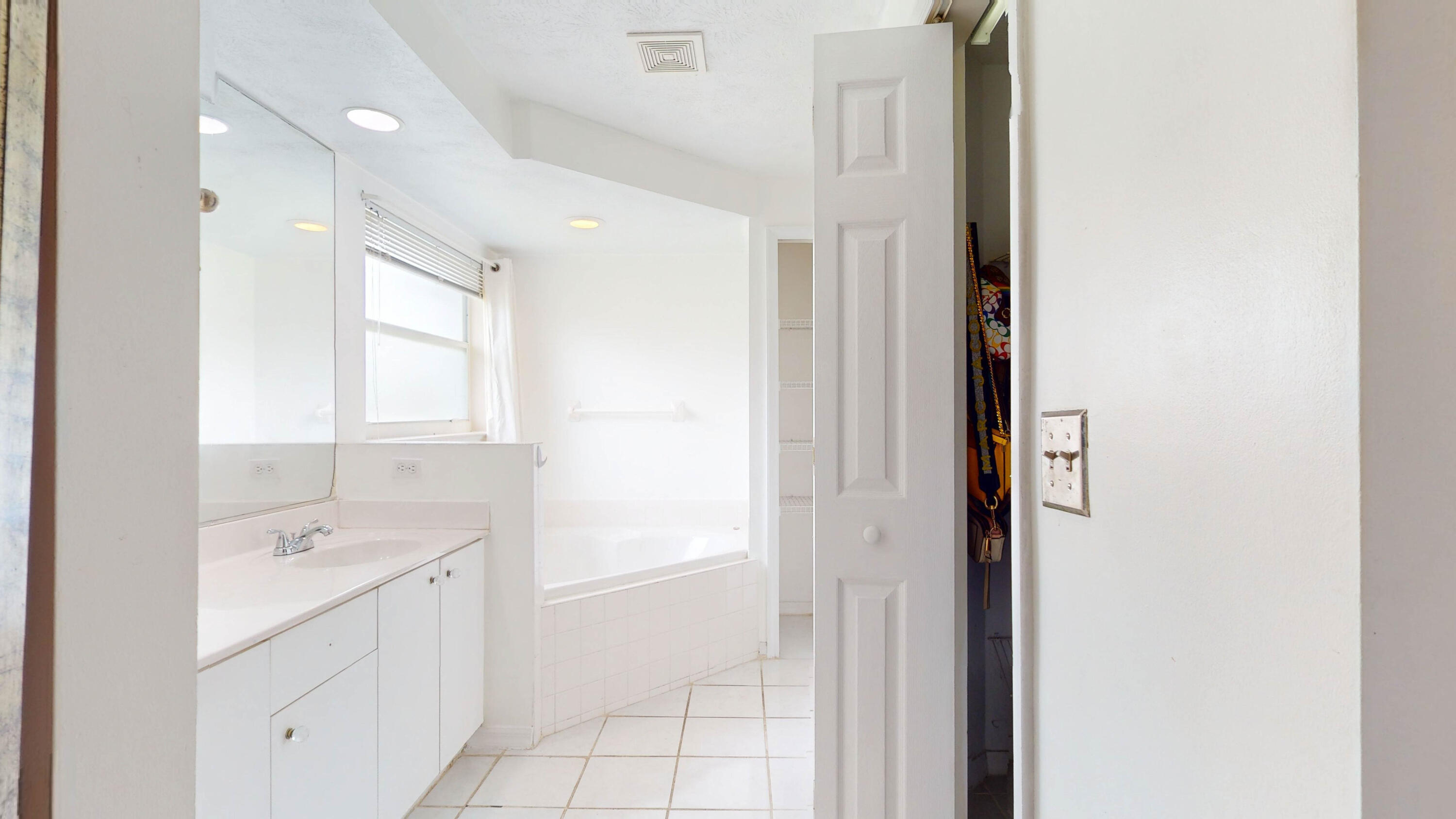 White bathroom with a mirrored vanity, tiled bathtub, and window with blinds.