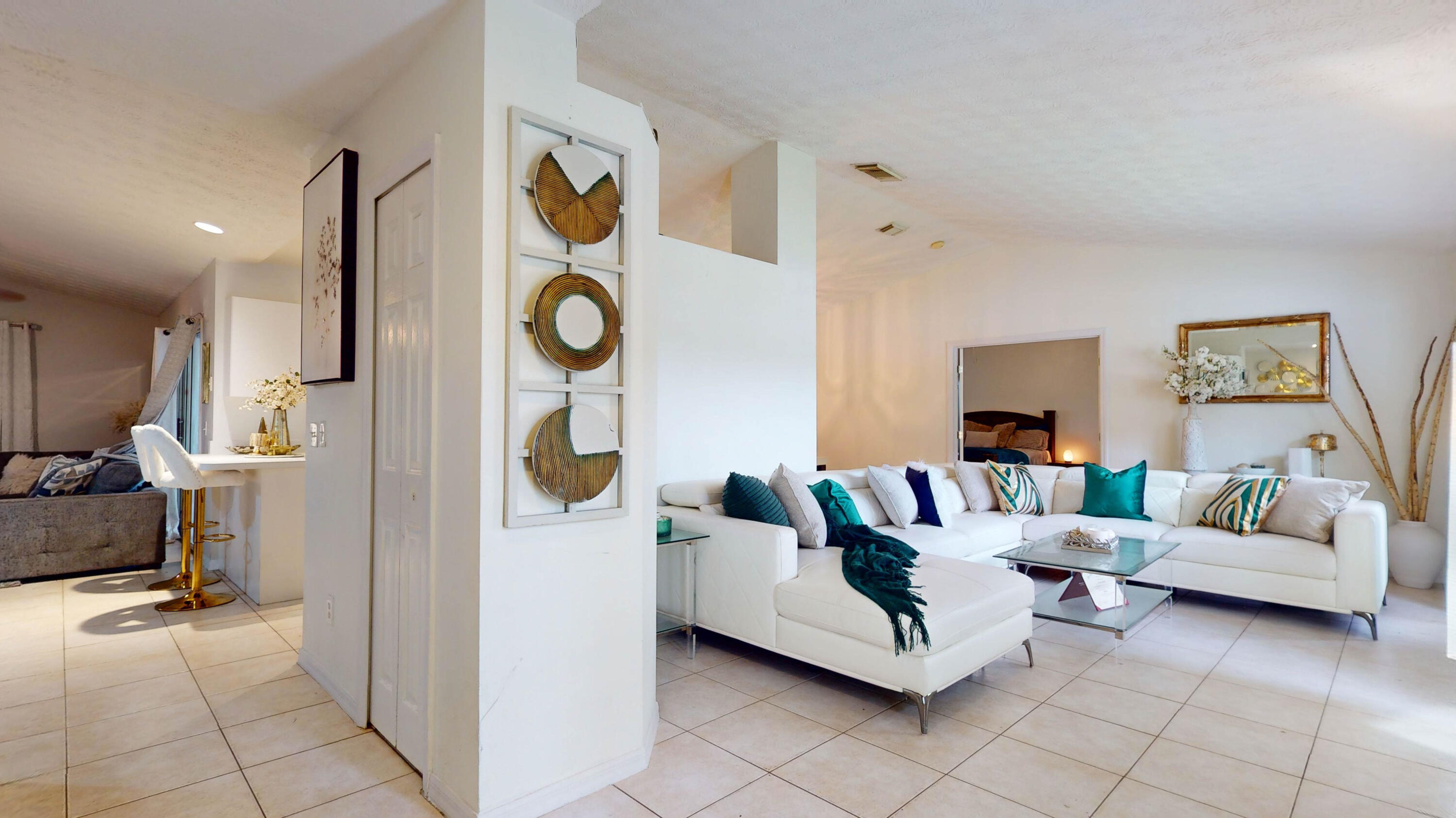 Staged living room with a white sectional, teal cushions, glass coffee table, and adjoining breakfast bar.