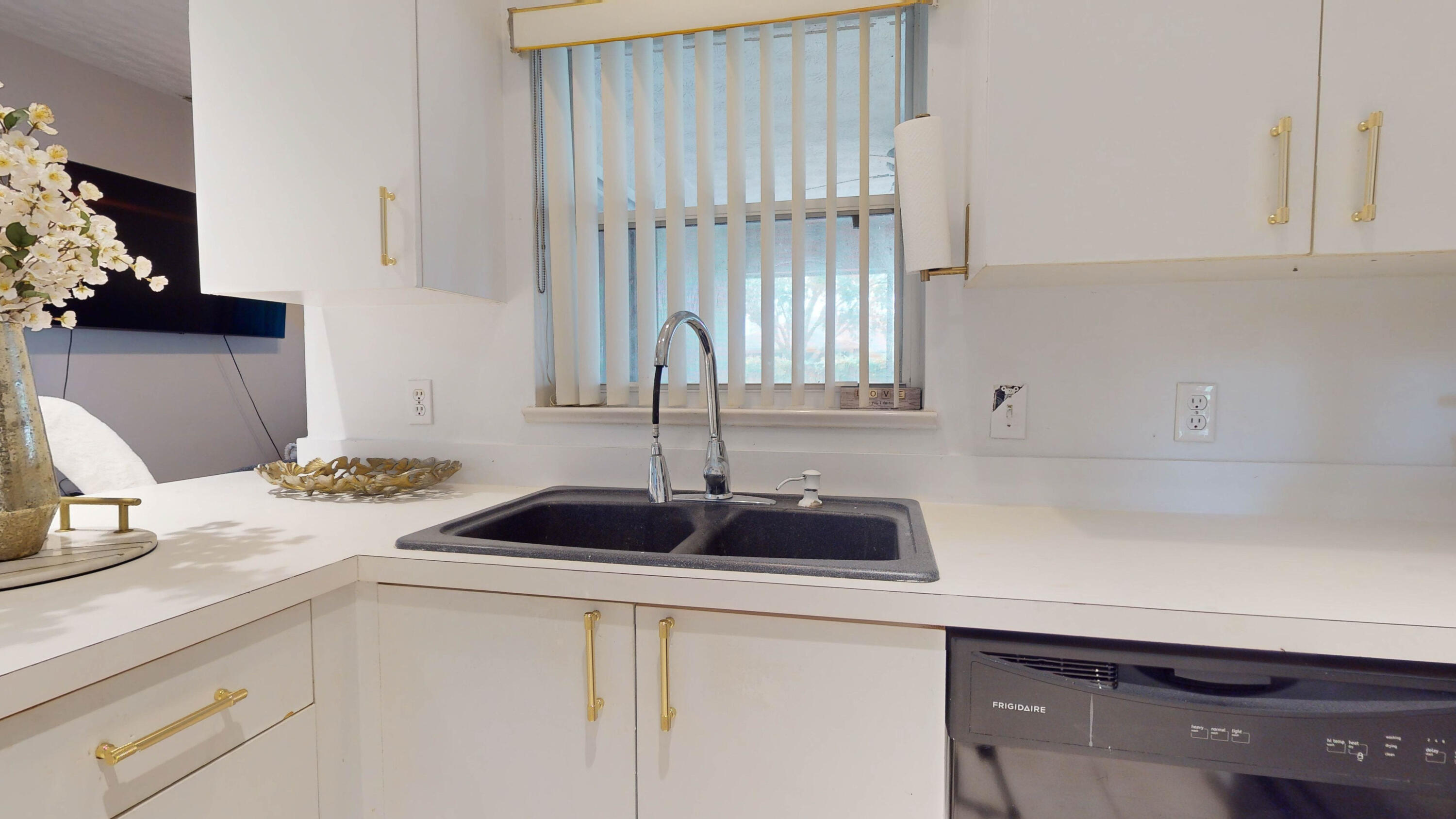Kitchen sink beneath a window, surrounded by white counters and cabinets with gold-tone hardware.