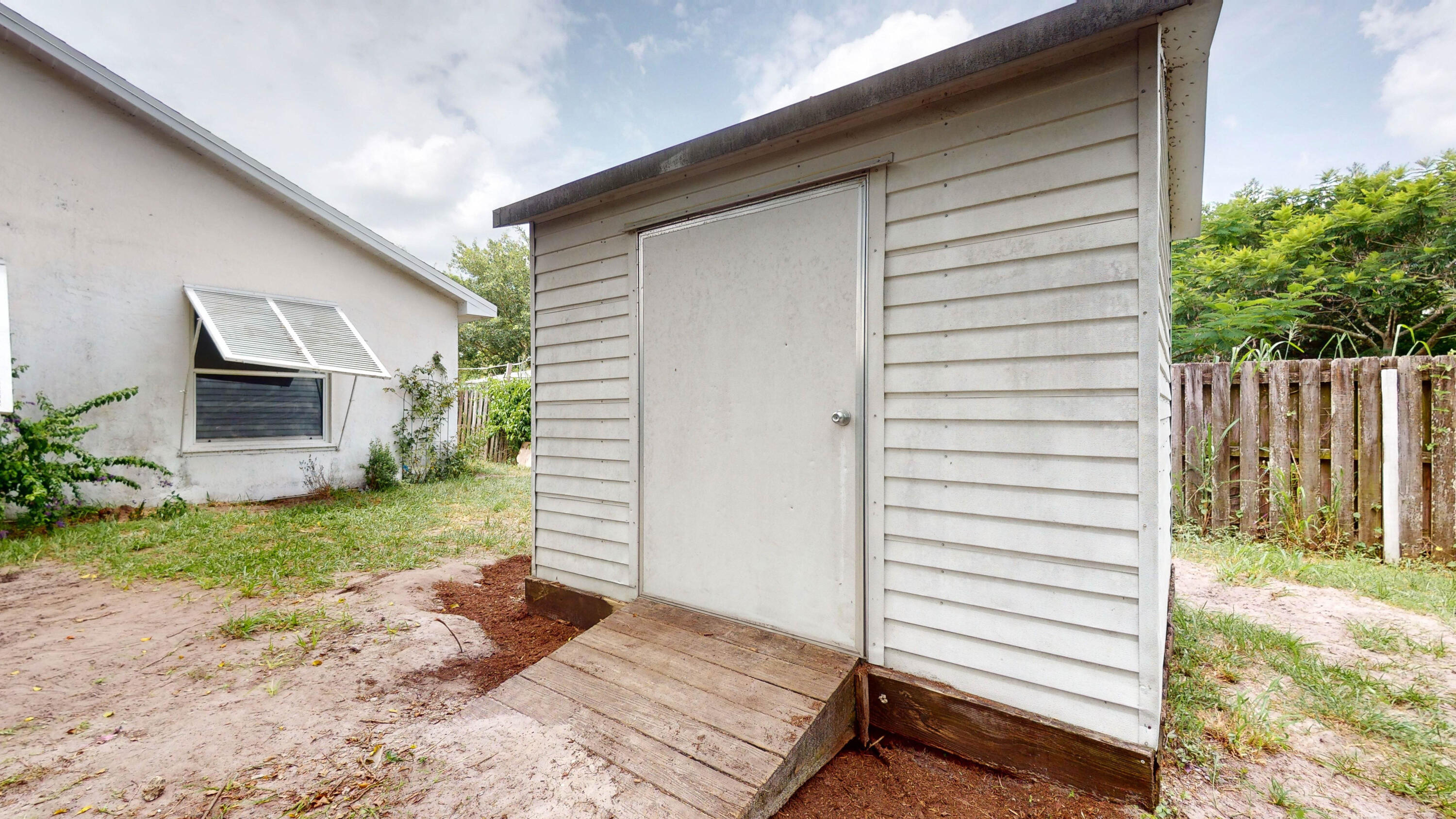 White-sided backyard shed with a wooden ramp, beside a neighboring building and fenced yard.