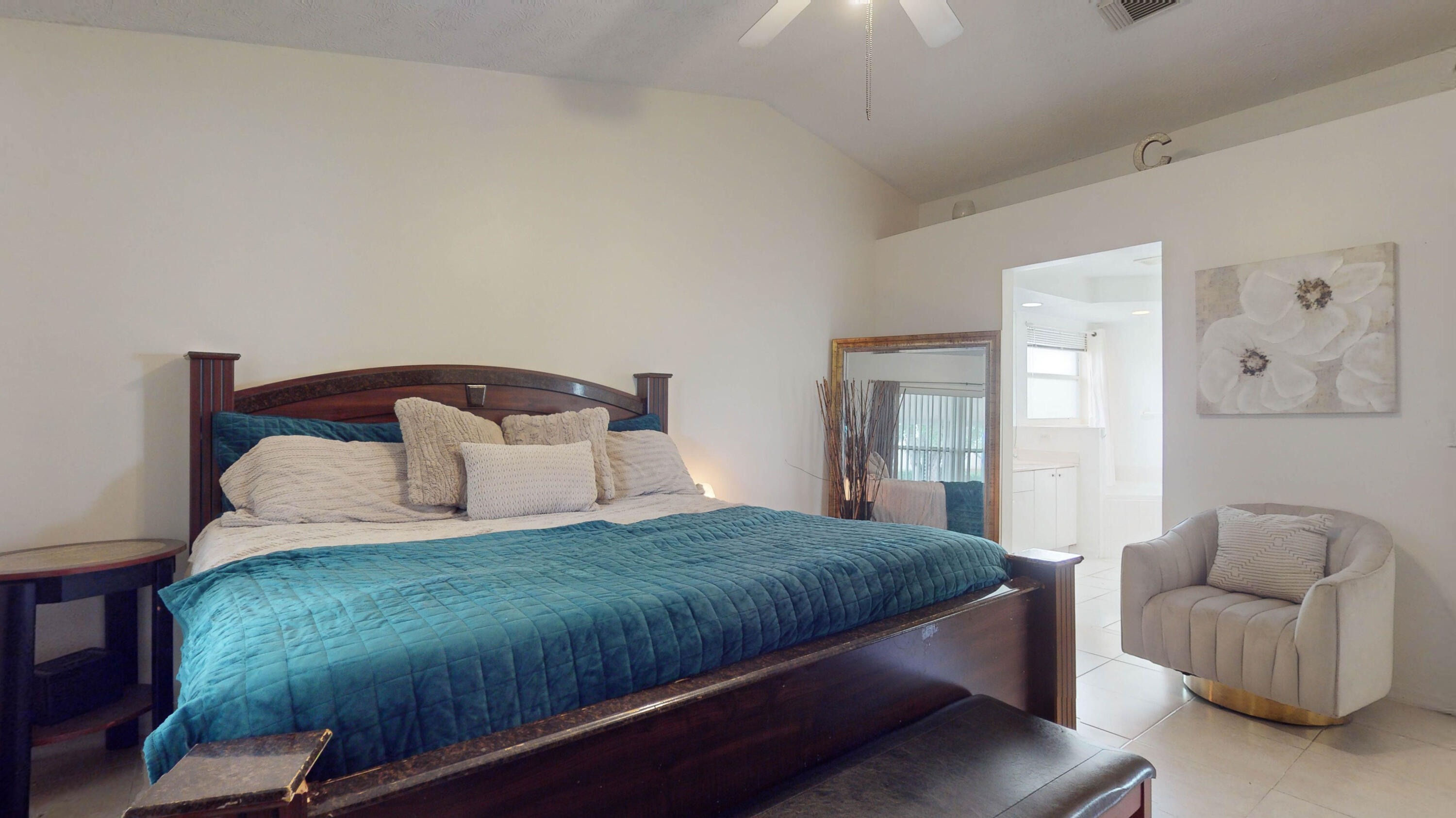 Bedroom with a dark wood bed, teal quilt, and beige armchair beside a bright adjoining room.