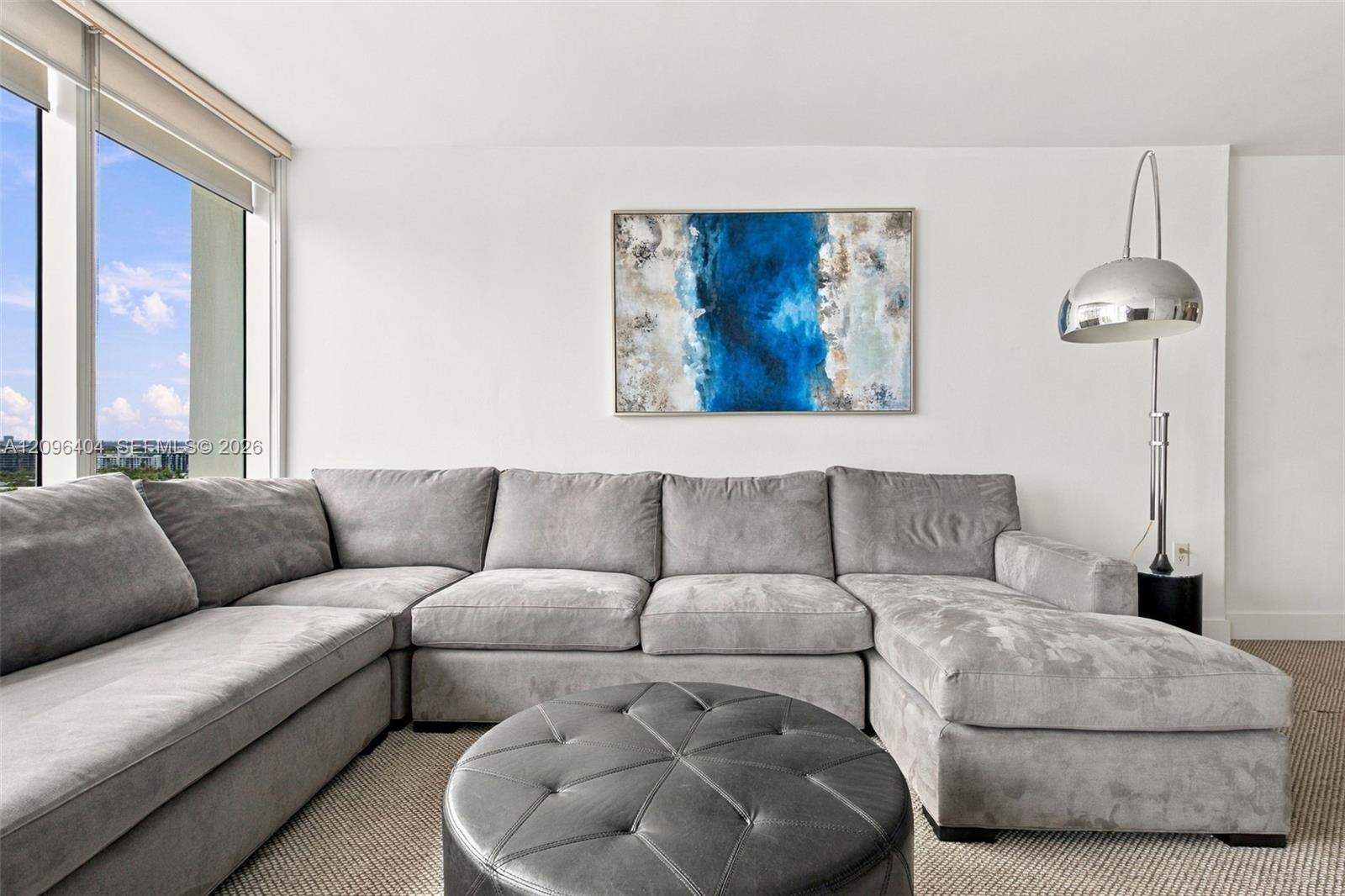 Living room with a gray sectional, round ottoman, blue abstract artwork, and tall windows.