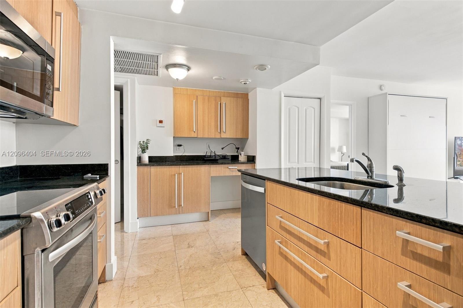 Kitchen with light wood cabinetry, dark countertops, a large island sink, and stainless steel appliances.