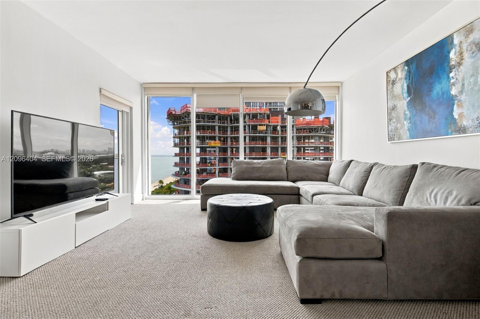 Condo living room with gray sectional and floor-to-ceiling windows overlooking water and nearby construction.
