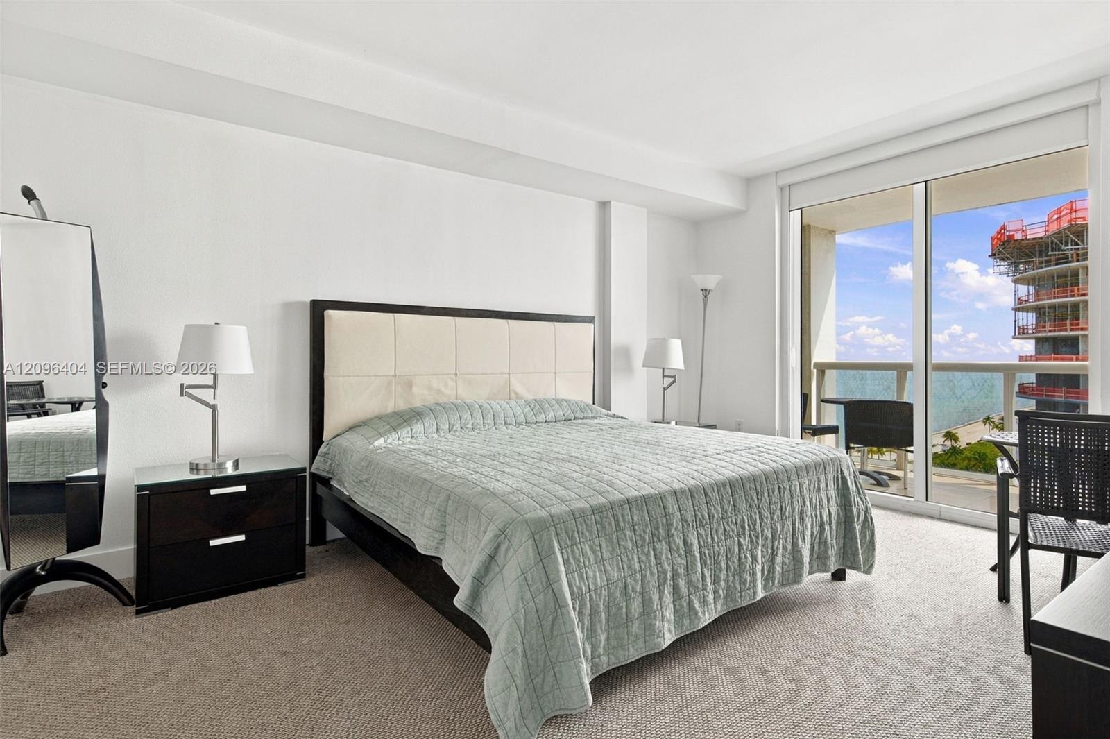Furnished bedroom with a large bed and balcony doors overlooking the water and a neighboring construction site.