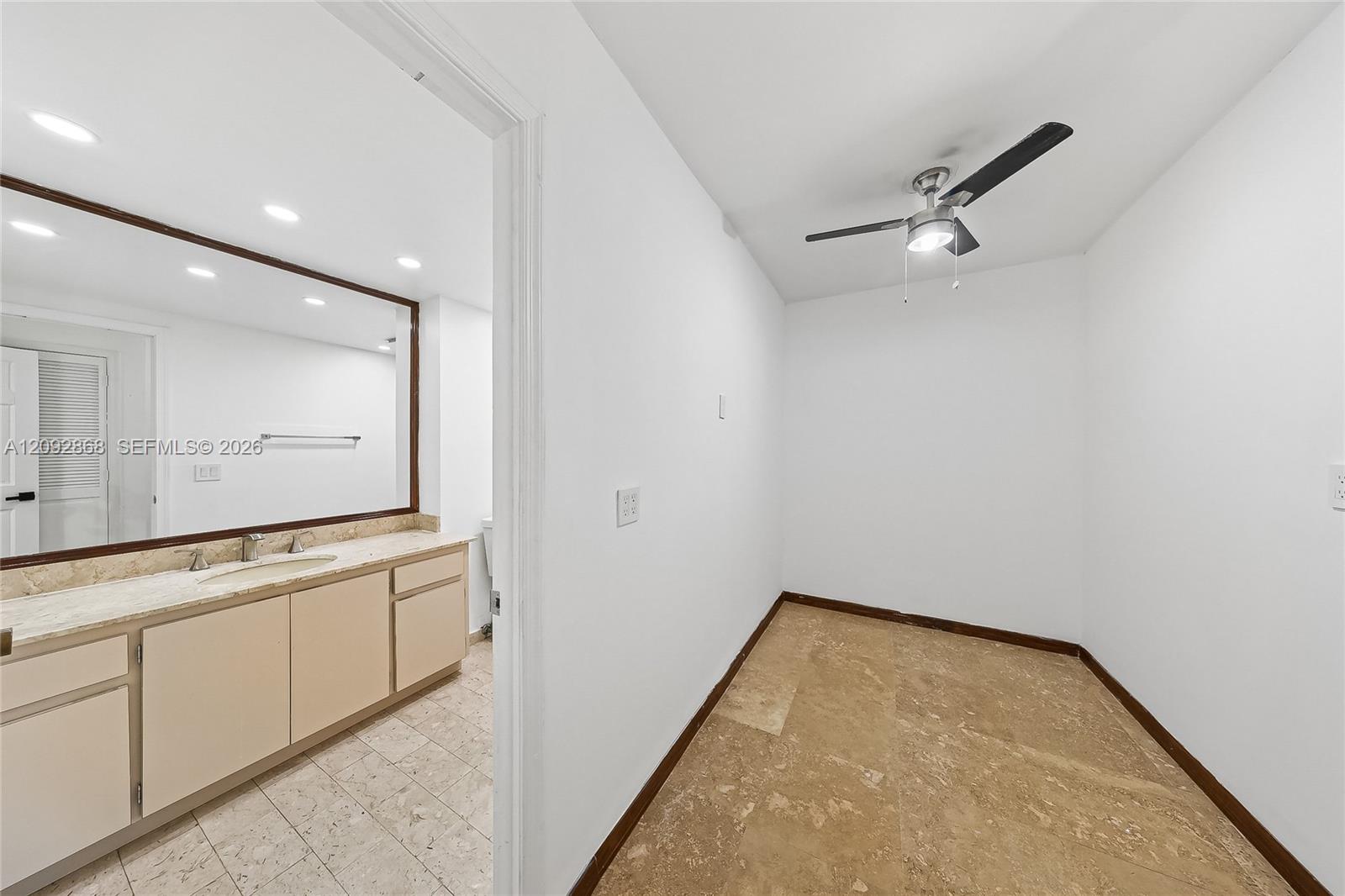 Vacant room with tan stone flooring and ceiling fan, beside a vanity and large mirror.
