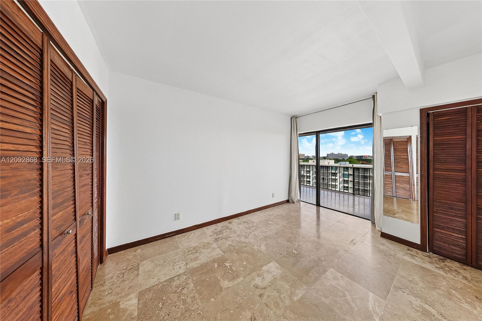 Empty bedroom with stone tile floors, louvered wood closets, and sliding glass doors to a balcony.