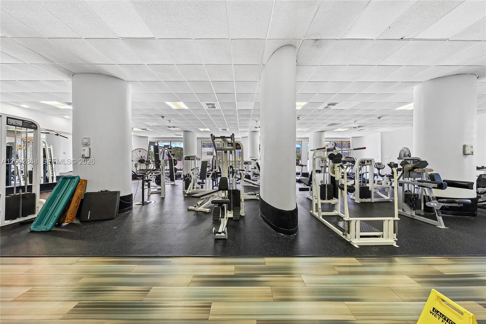 Fitness center with rows of exercise machines between large white columns.