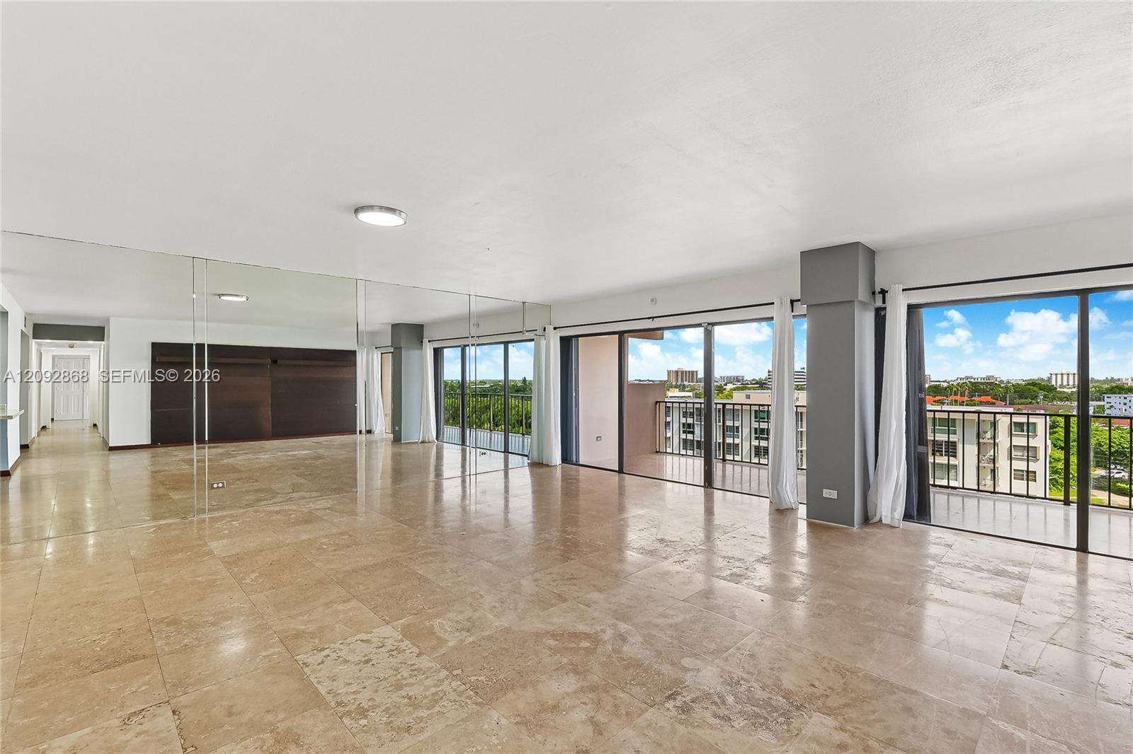Vacant living room with tan stone tile, mirrored wall, and balcony windows overlooking buildings and greenery.