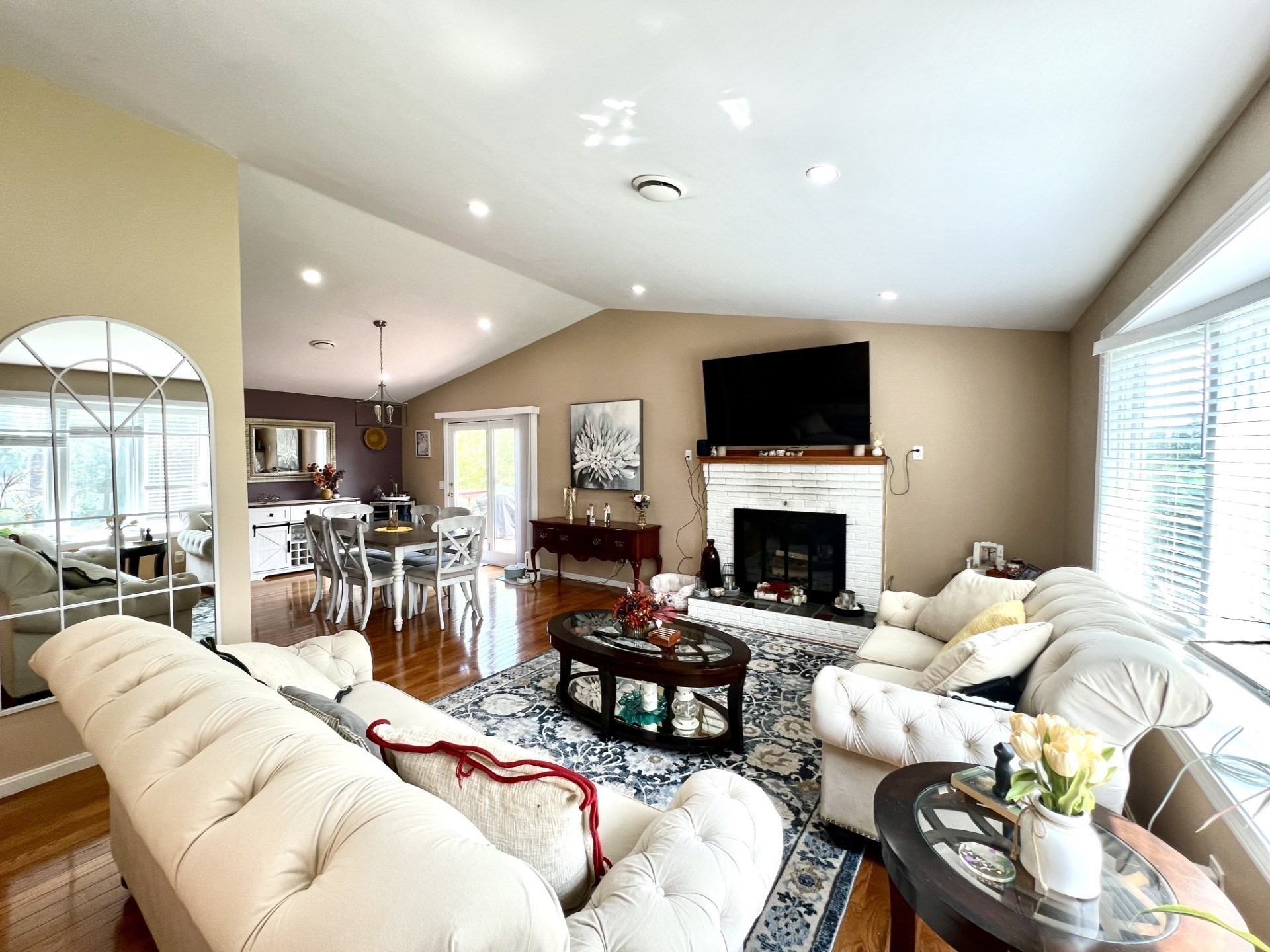 Furnished living room with white sofas, a white brick fireplace, vaulted ceiling, and adjoining dining area.