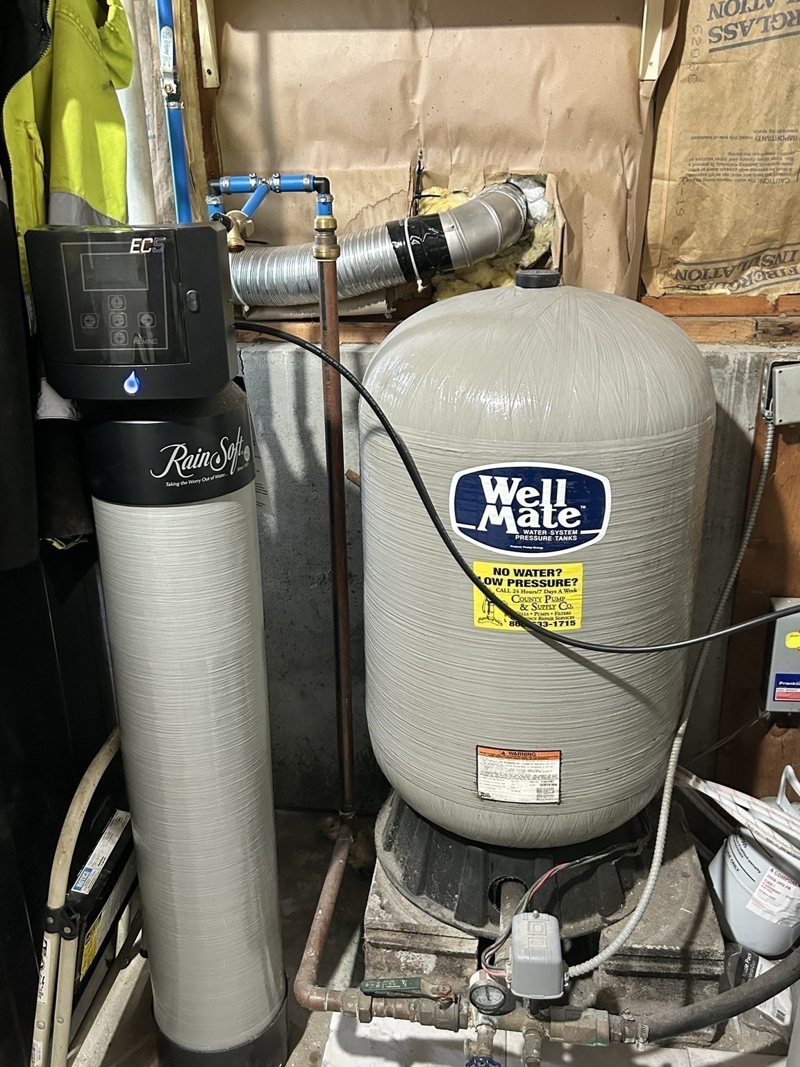 Water softener and large pressure tank with exposed plumbing in a home's utility area.