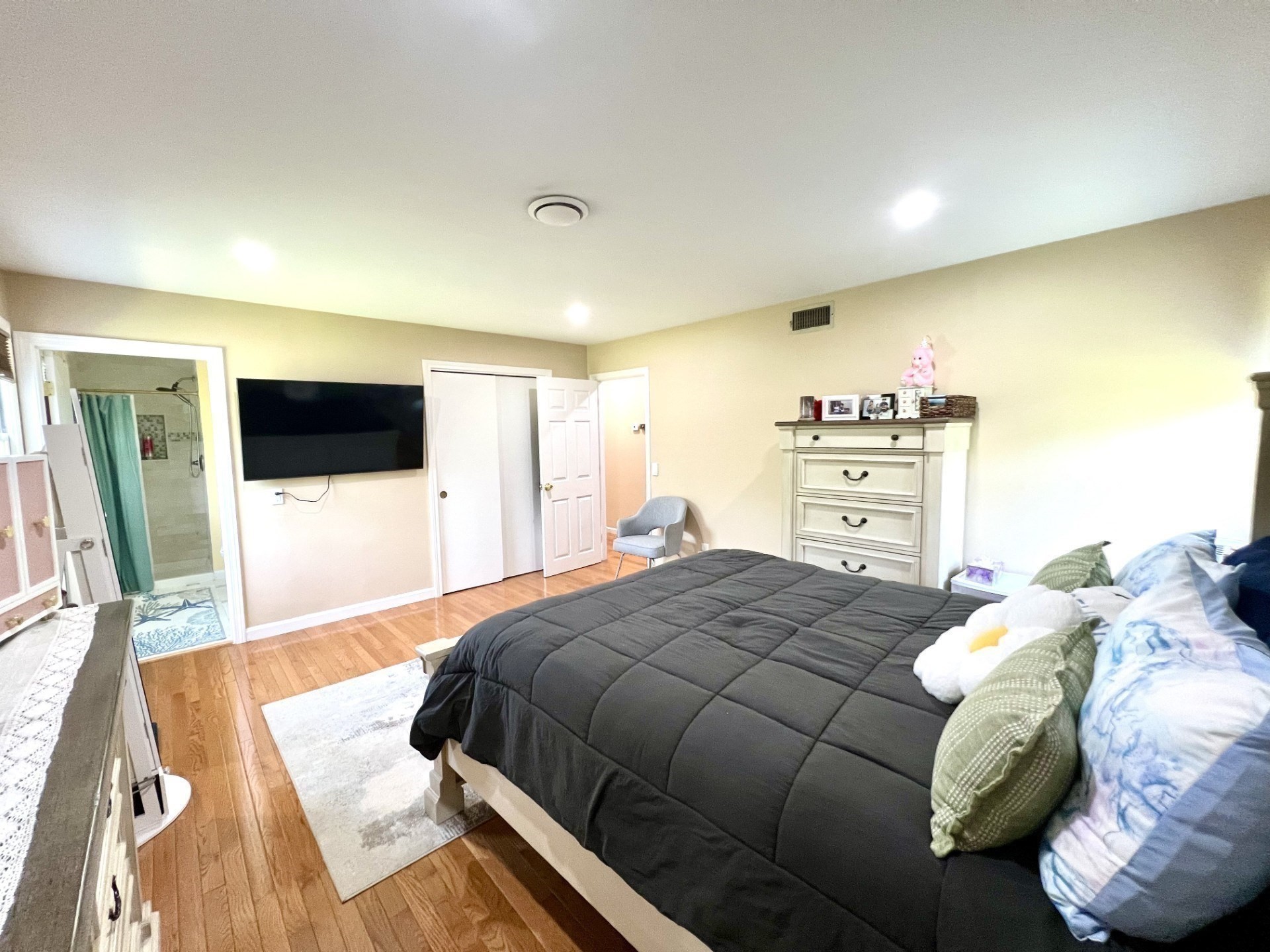 Bedroom with a dark-quilted bed, hardwood floors, wall-mounted TV, and adjoining bathroom.