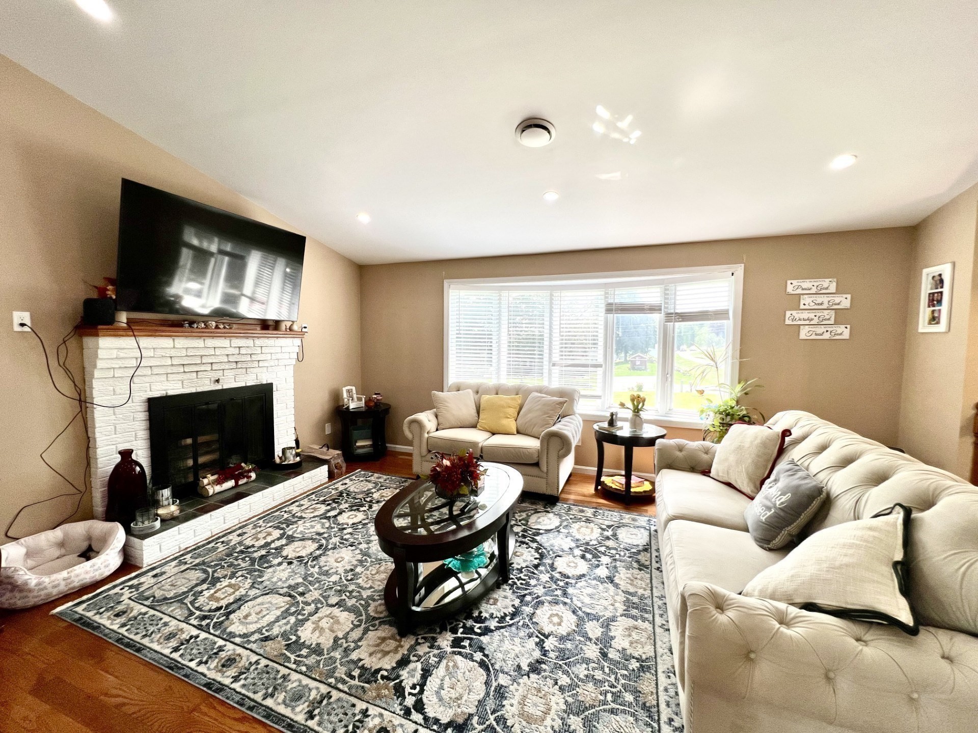 Furnished living room with white brick fireplace, large window, cream sofas, and patterned rug.