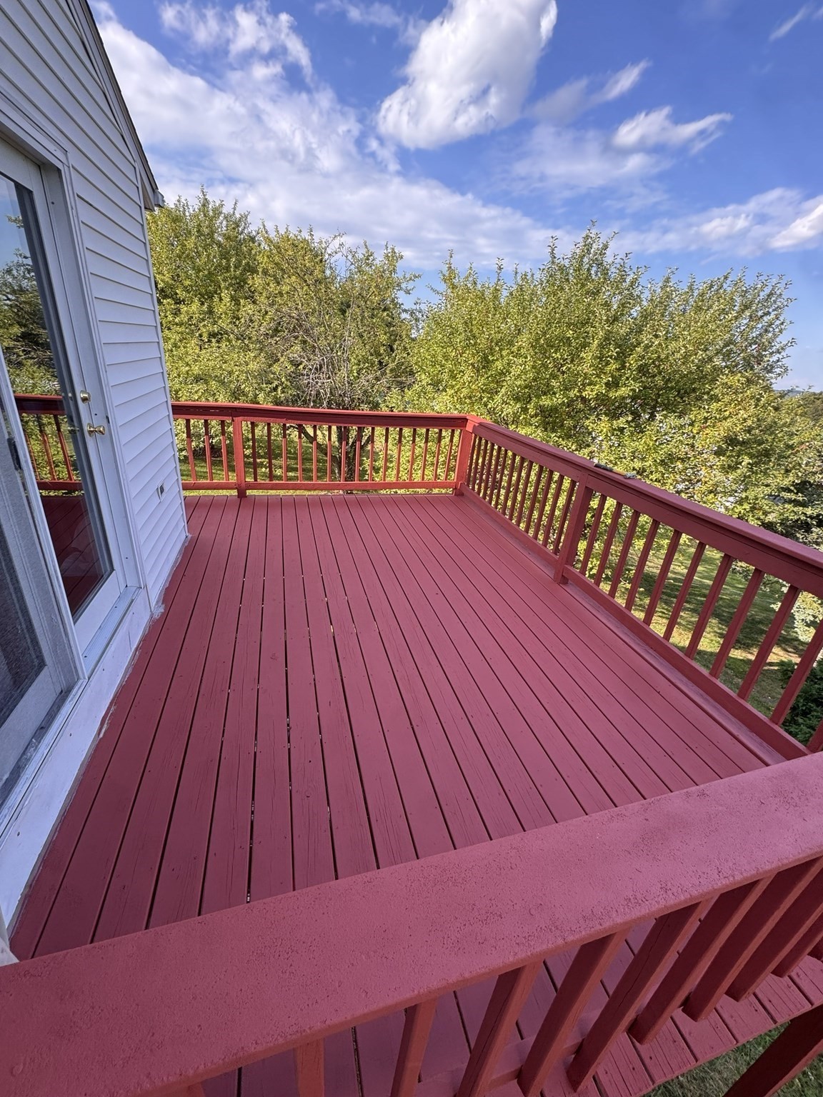 Red-painted wooden deck with matching railings beside a white-sided house, overlooking leafy trees.