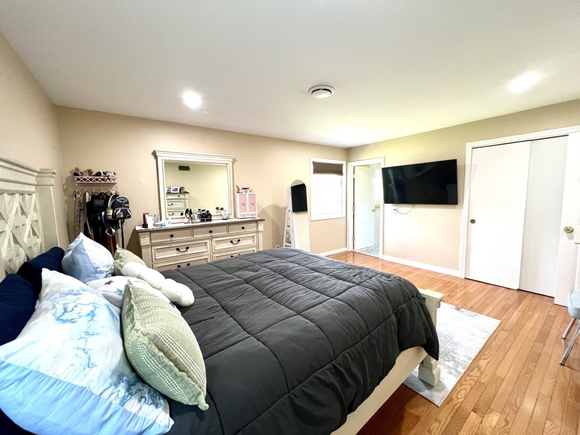 Furnished bedroom with a large gray-quilted bed, dresser and mirror, wall-mounted TV, and hardwood floors.