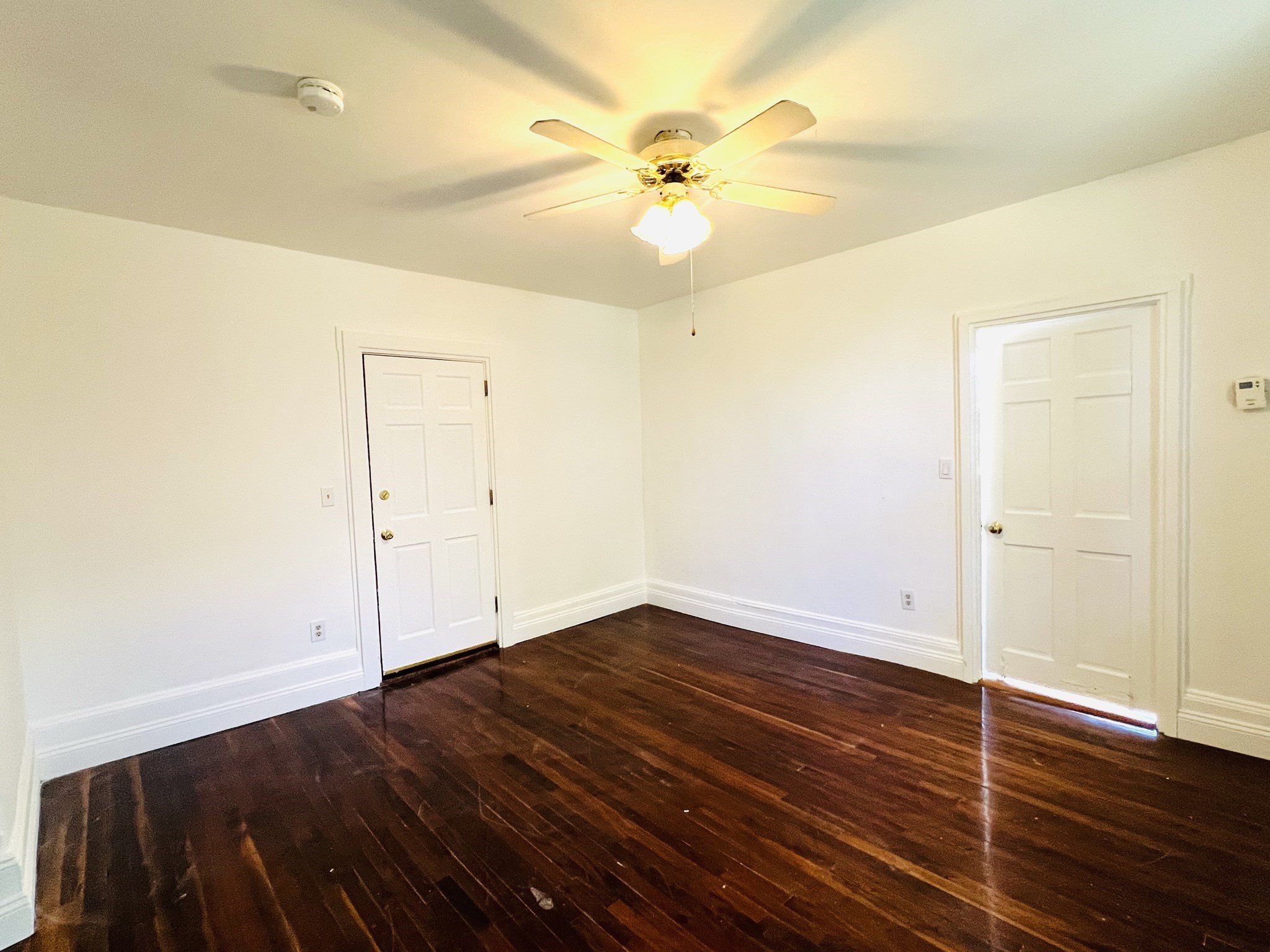 Empty room with dark hardwood floors, white paneled doors, and a lit ceiling fan.