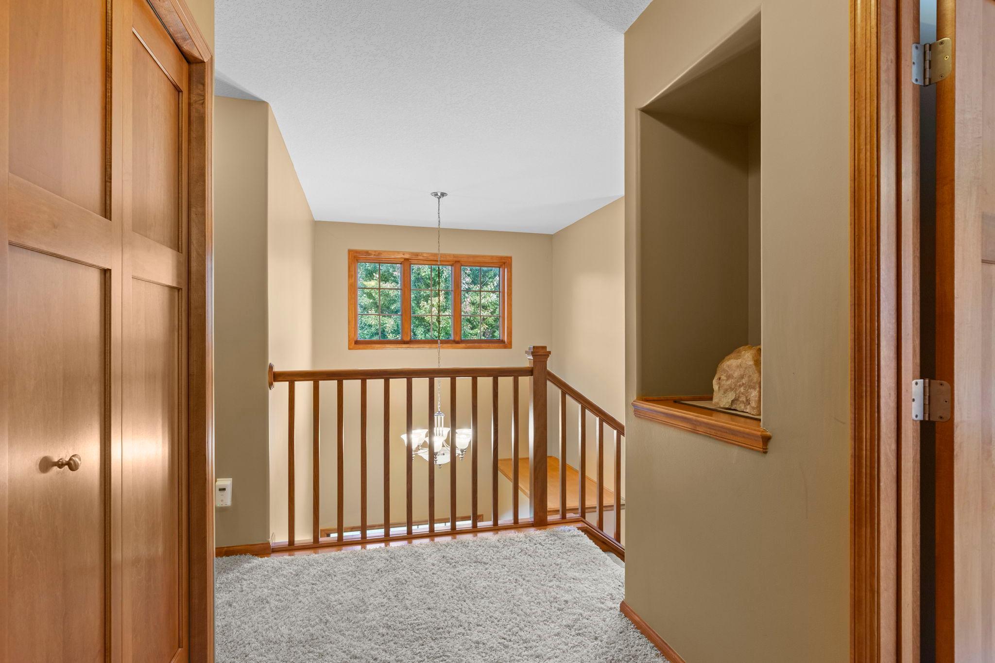 Upstairs landing with wood stair railing, carpet, a trimmed window, and hanging chandelier.