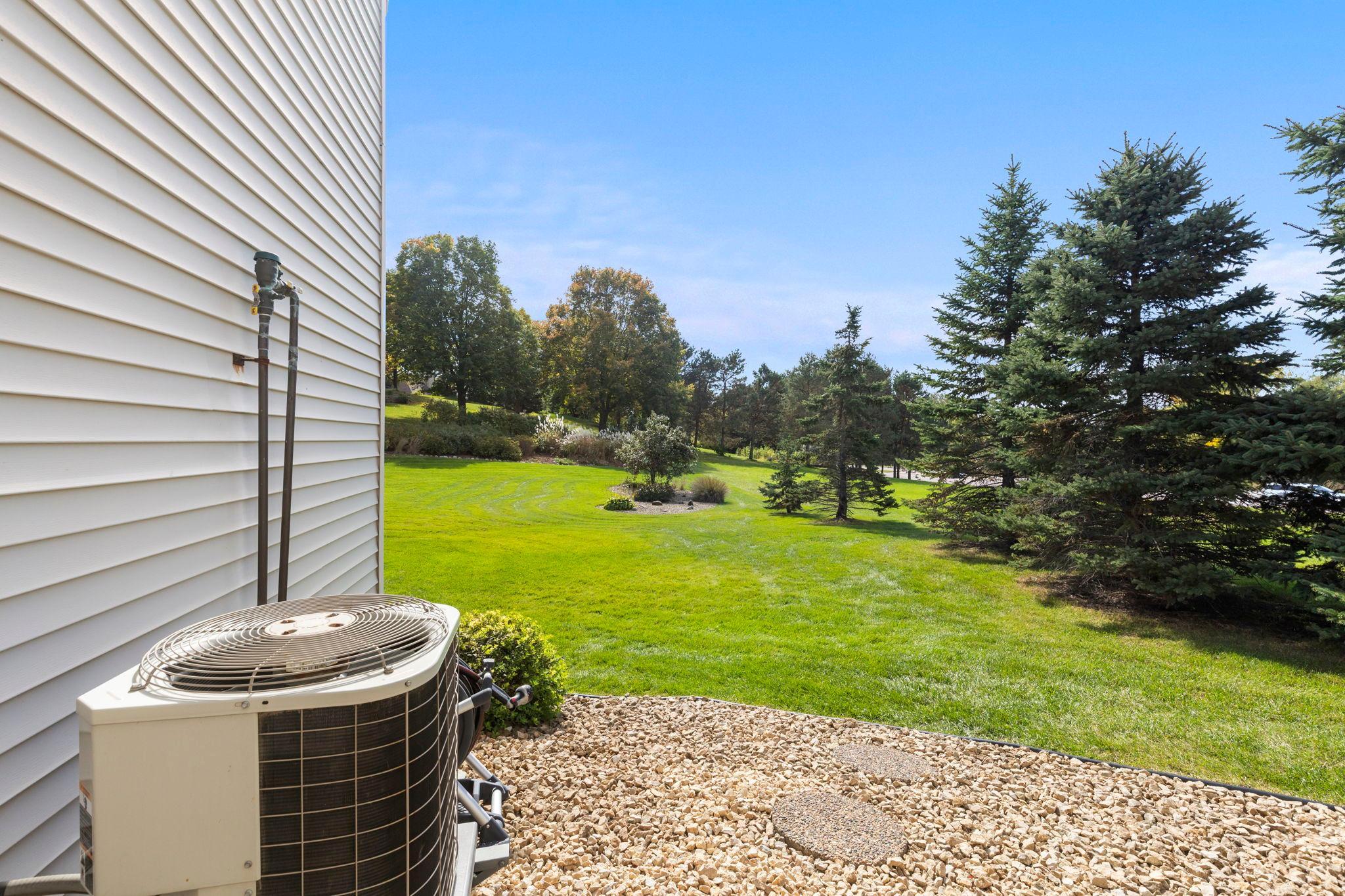 Side yard with a broad lawn, evergreen trees, and an outdoor air-conditioning unit beside the house.