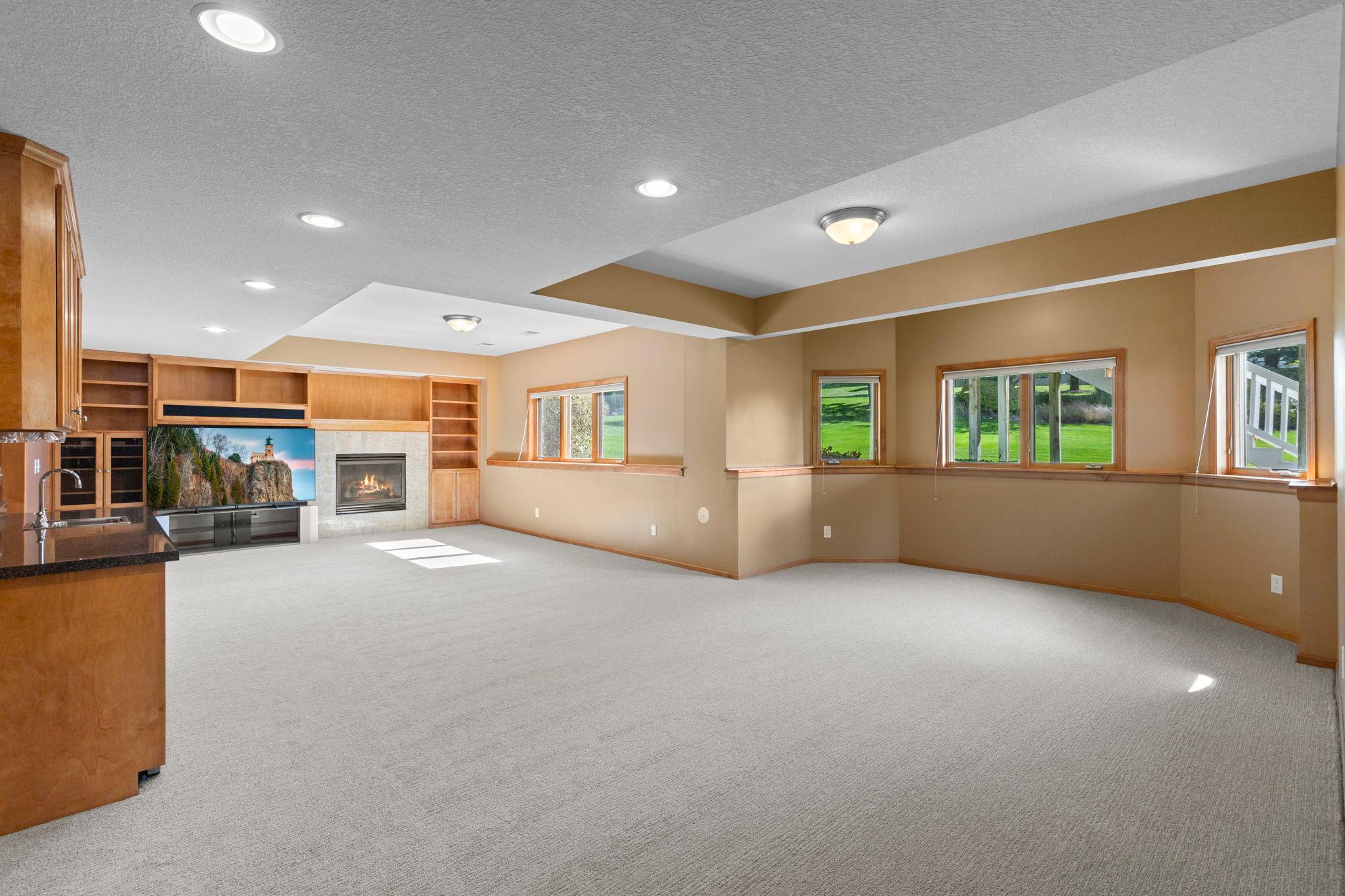 Carpeted living room with built-in wood shelving, a stone fireplace, and windows overlooking a lawn.