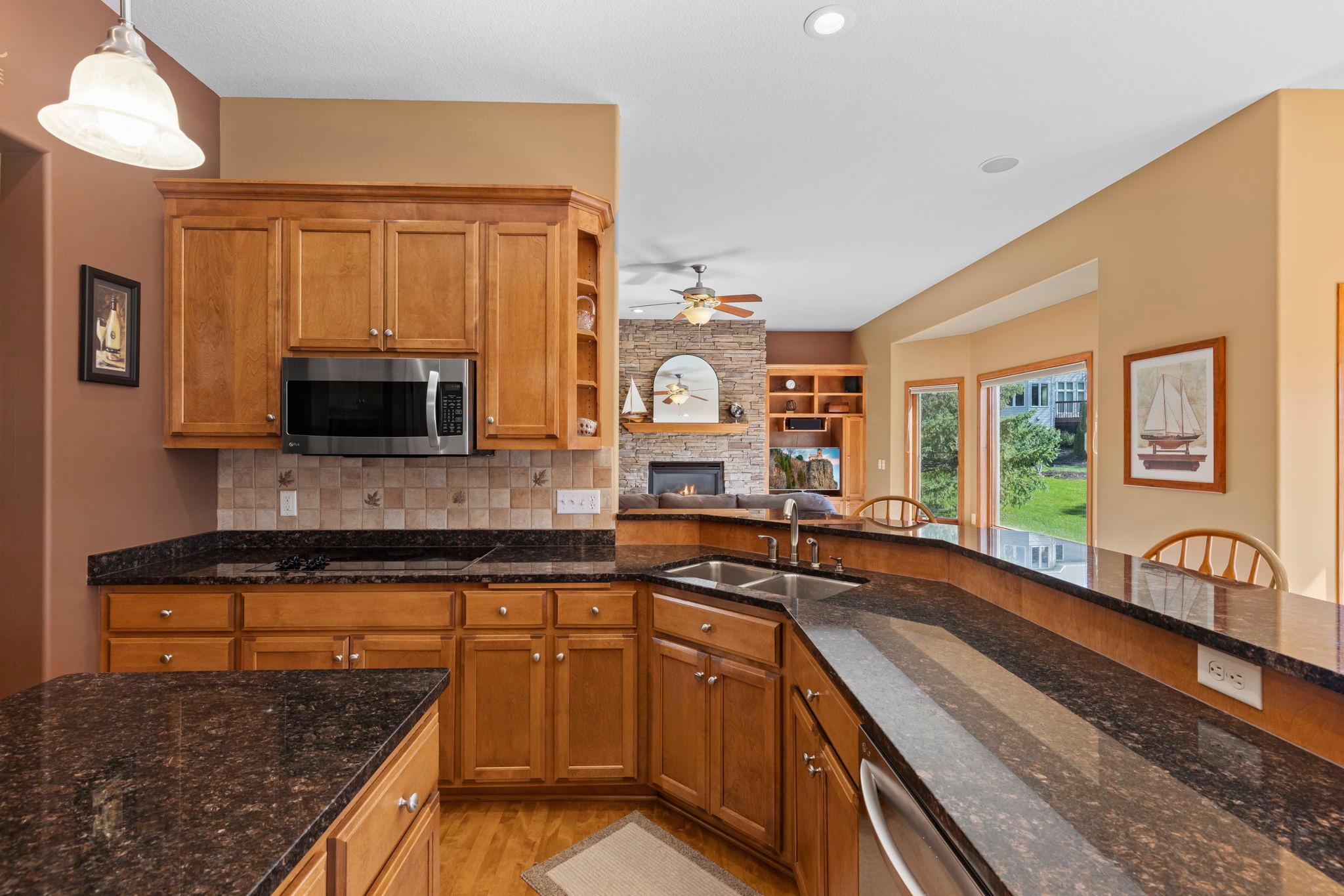 Kitchen with warm wood cabinets and granite counters, opening to a living area with a stone fireplace.