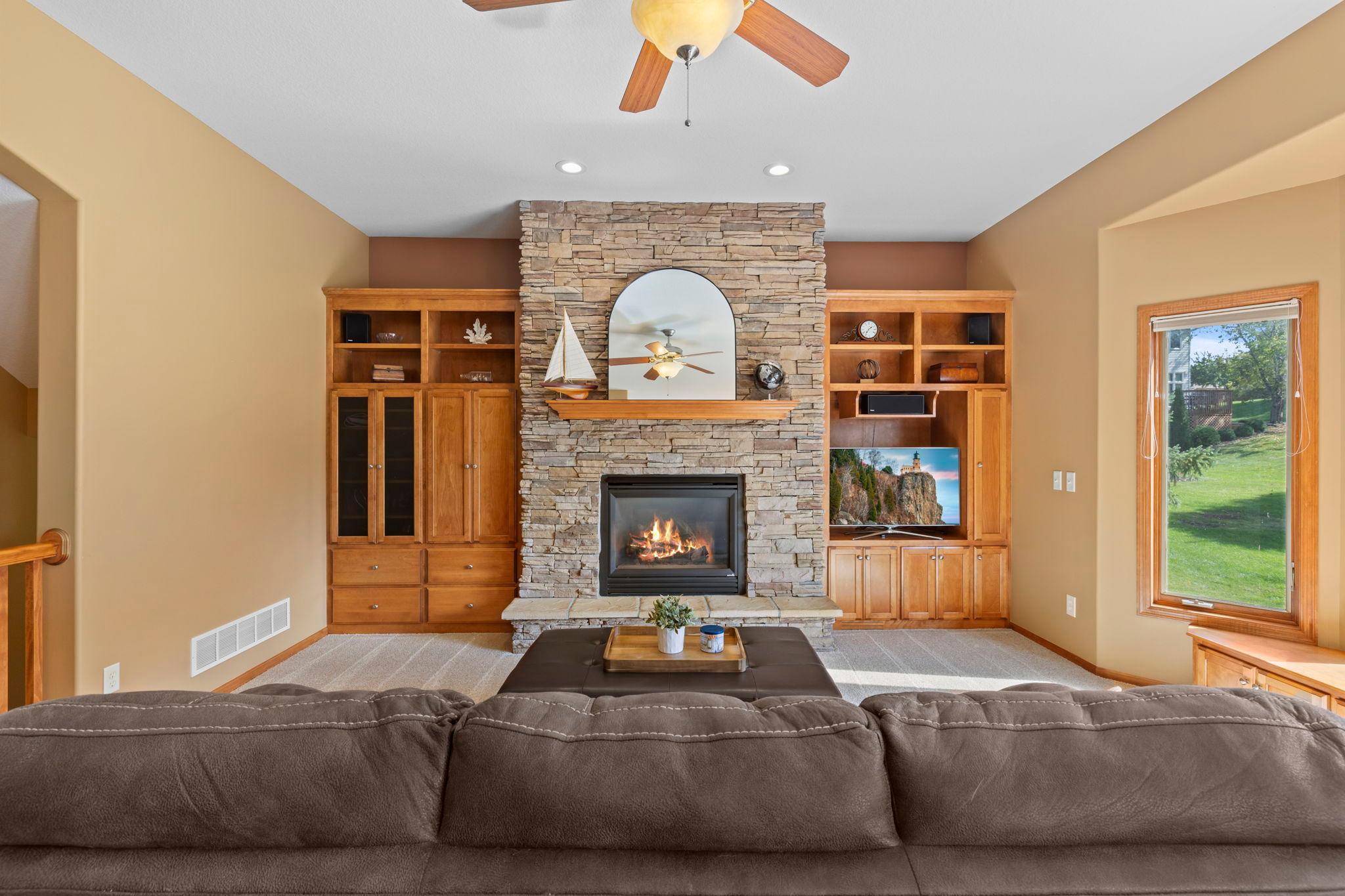 Living room with a stone fireplace, built-in wooden cabinets, and a large window overlooking the lawn.