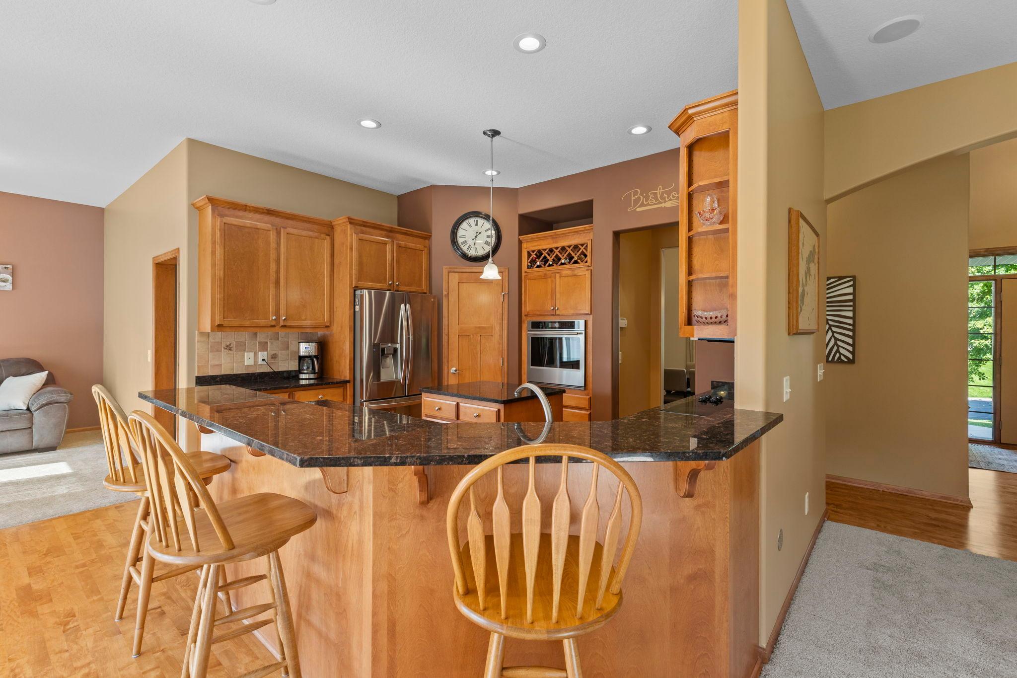 Kitchen with a curved granite breakfast bar, wood cabinetry, stainless steel appliances, and wooden stools.