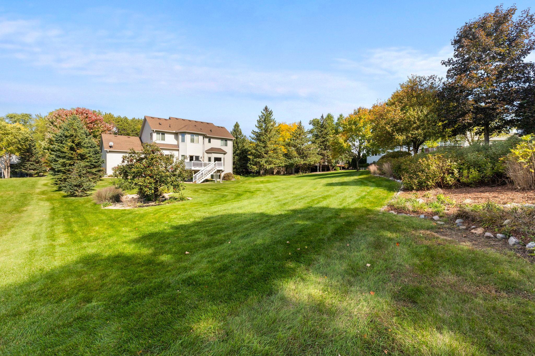 Two-story home's backyard with a wide lawn, mature trees, and a raised deck with stairs.