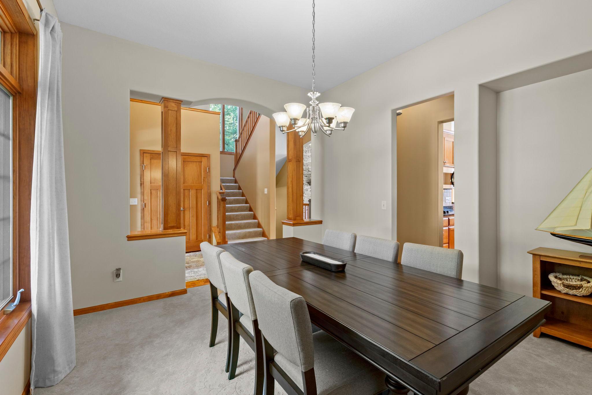 Dining room with a long dark wood table, gray chairs, and a chandelier beside a carpeted staircase.