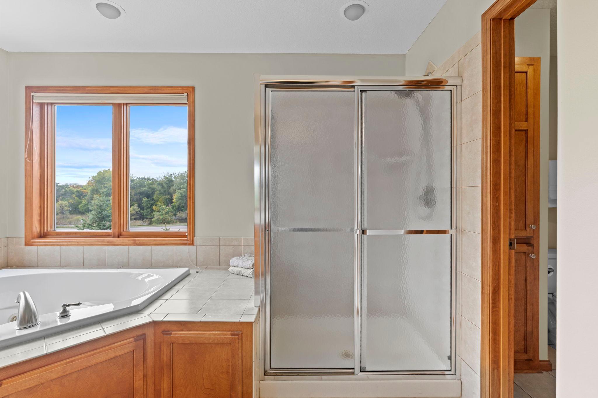 Bathroom with a corner soaking tub, glass-enclosed shower, and wood-trimmed window overlooking trees.
