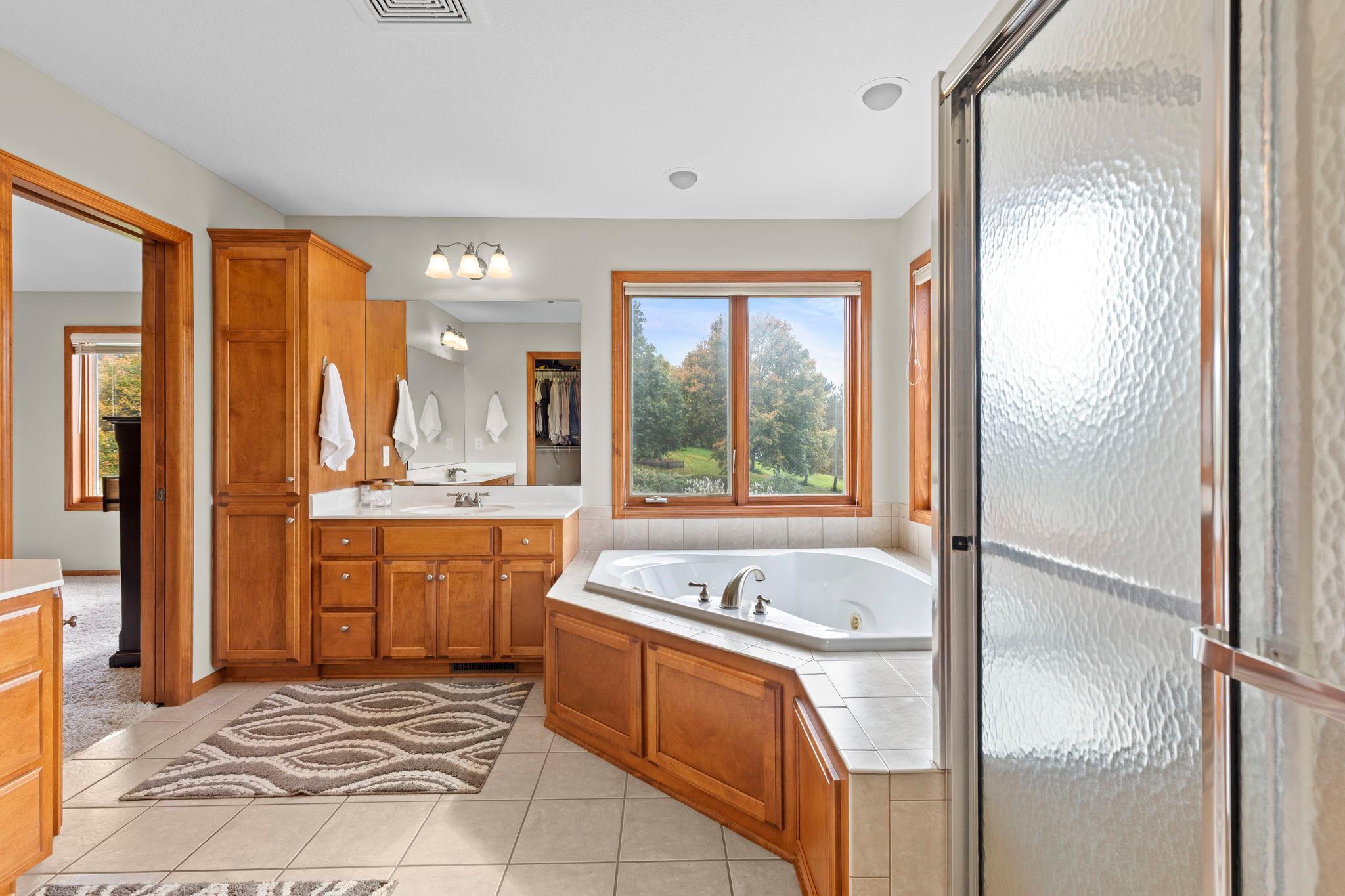 Primary bathroom with a corner soaking tub, warm wood cabinetry, and a large window overlooking trees.