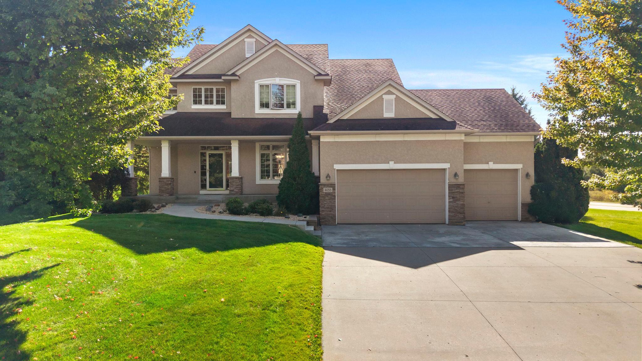 Two-story beige stucco home with a covered porch, three-car garage, and broad driveway.