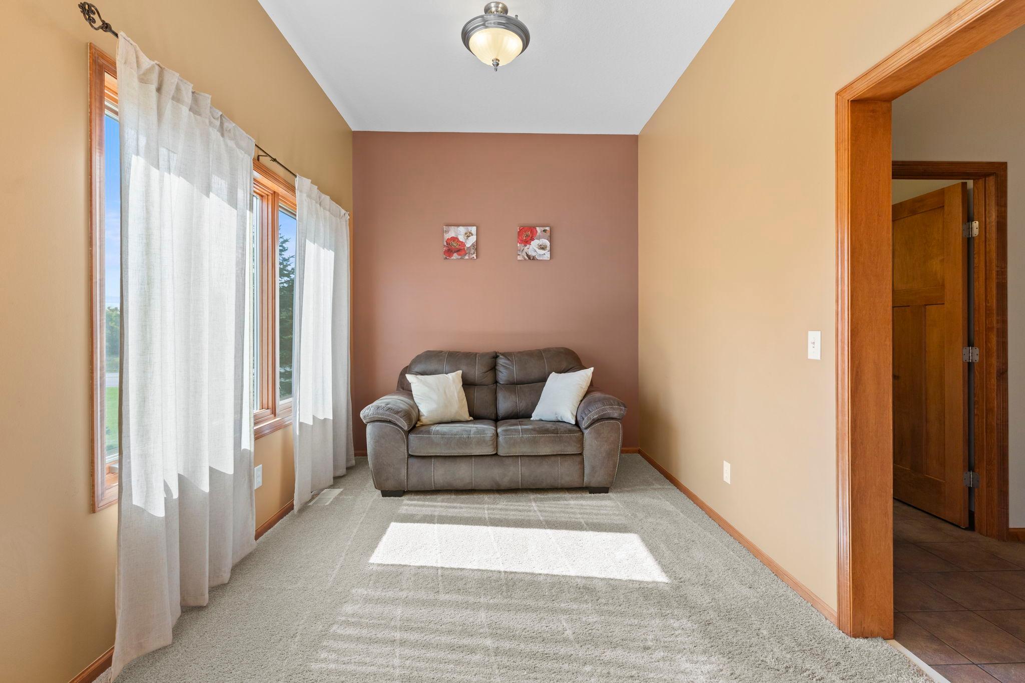 Sitting room with brown loveseat, tall curtained windows, and sunlight on the carpet.