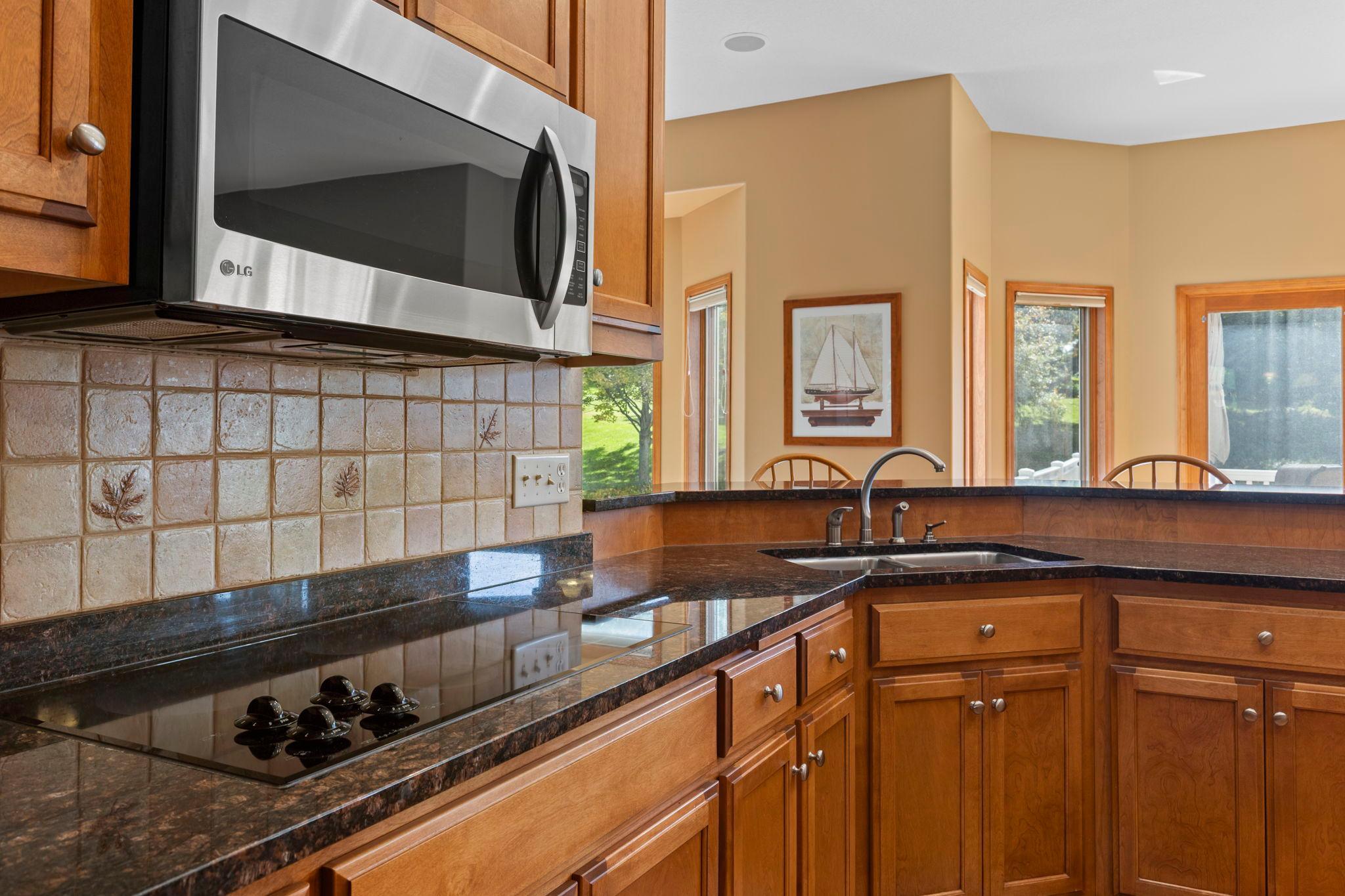 Kitchen with wood cabinetry, dark granite counters, stainless microwave, and a sink beside bright windows.