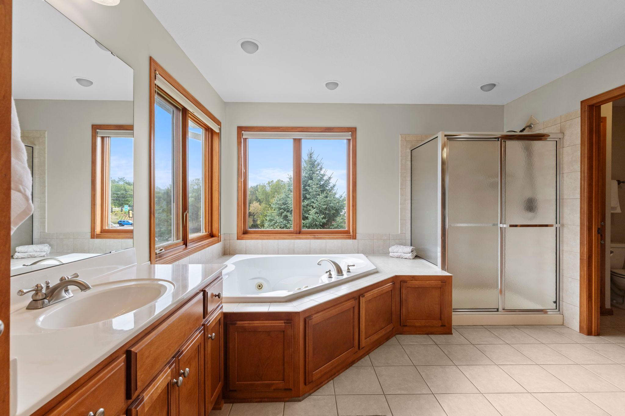 Primary bathroom with a corner whirlpool tub, wood cabinetry, and a glass-enclosed shower beside large windows.