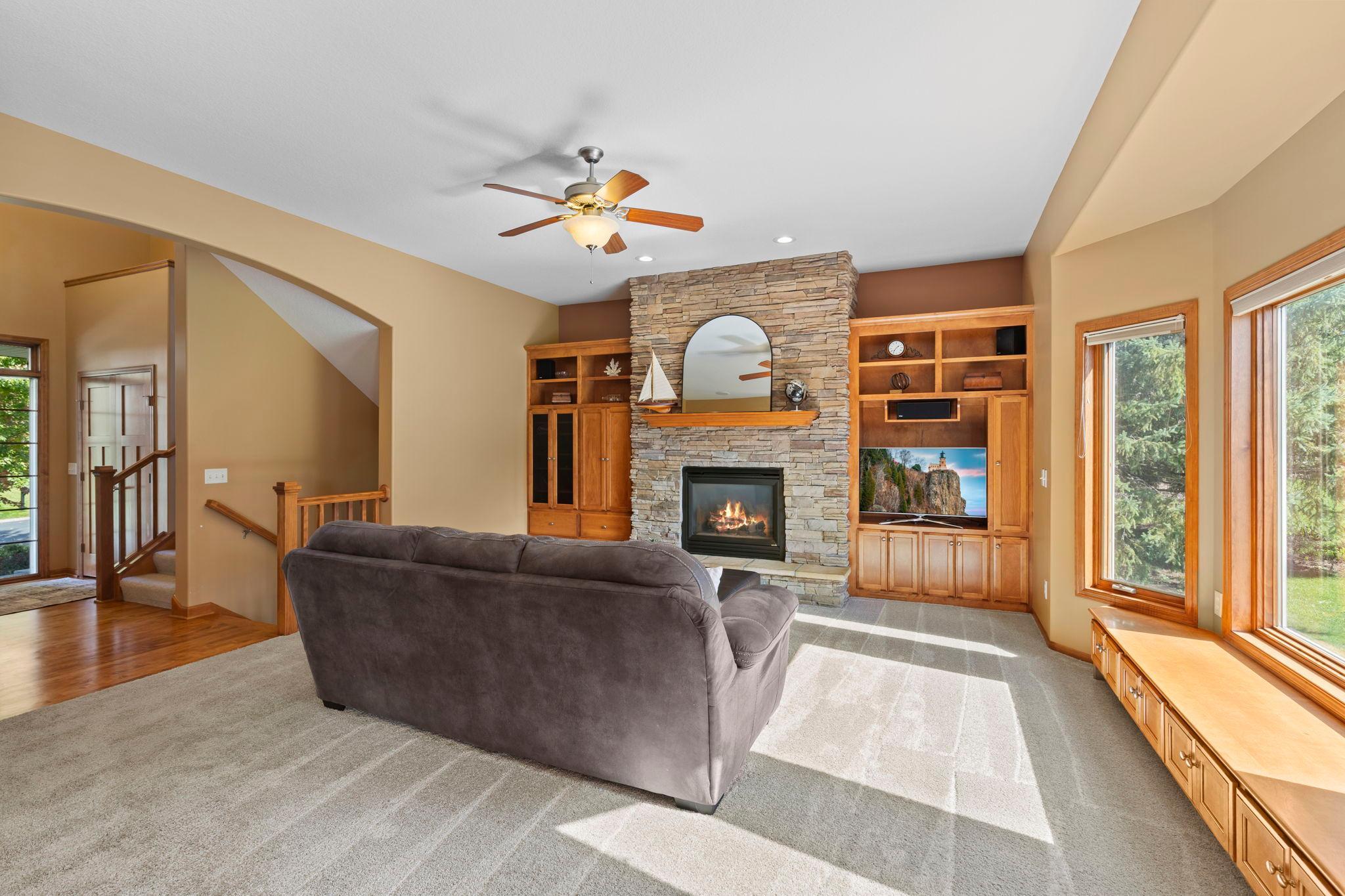 Living room with a stone fireplace, built-in wooden cabinetry, and large sunlit windows.